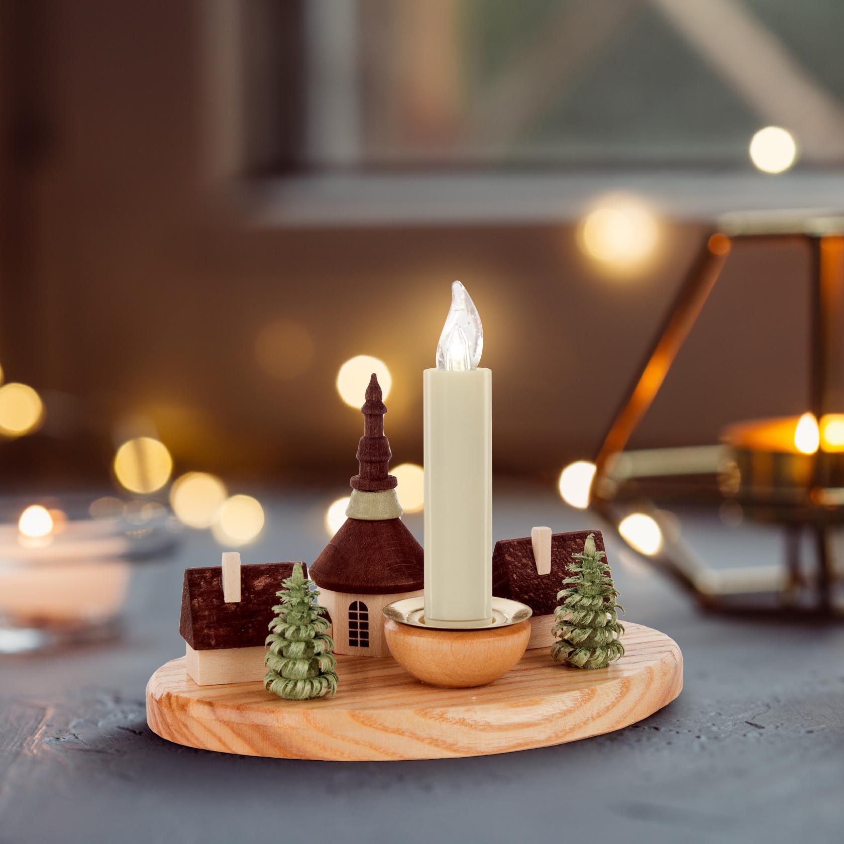 A wooden candle holder depicting a miniature village scene with two houses, trees, and a church from Seiffen. A candle is in the middle.