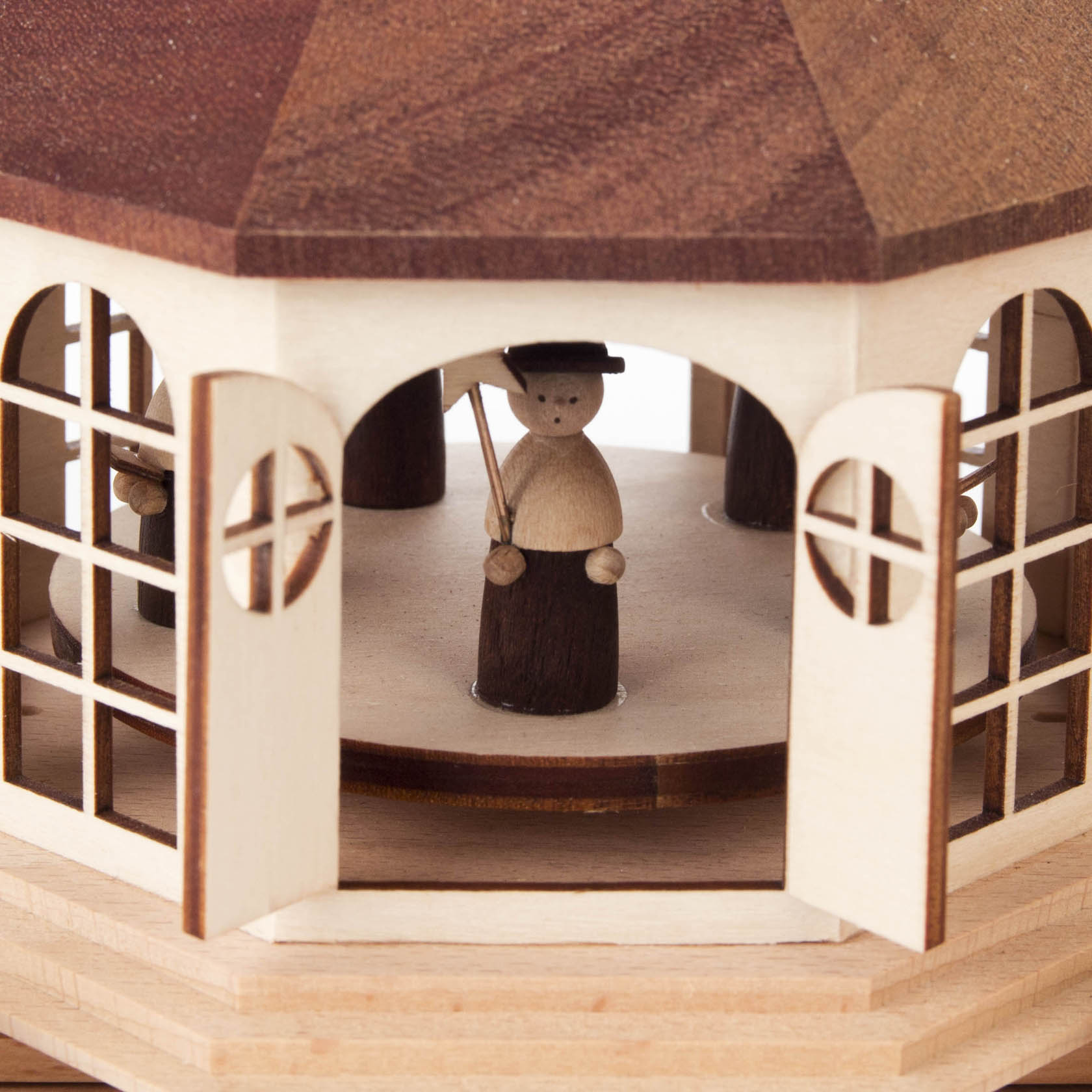 Detailaufnahme einer Holzbogenkrippe mit einer kleinen Holzfigur einer Kurrende, gekleidet in dunklem Holzton und einem runden Hut, im Innenraum.