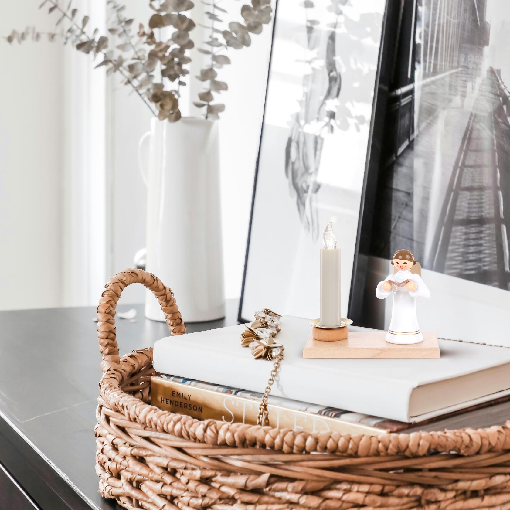 A decorative candle holder with a standing angel figurine and a white candle on a wooden base, placed on a book in a woven basket.