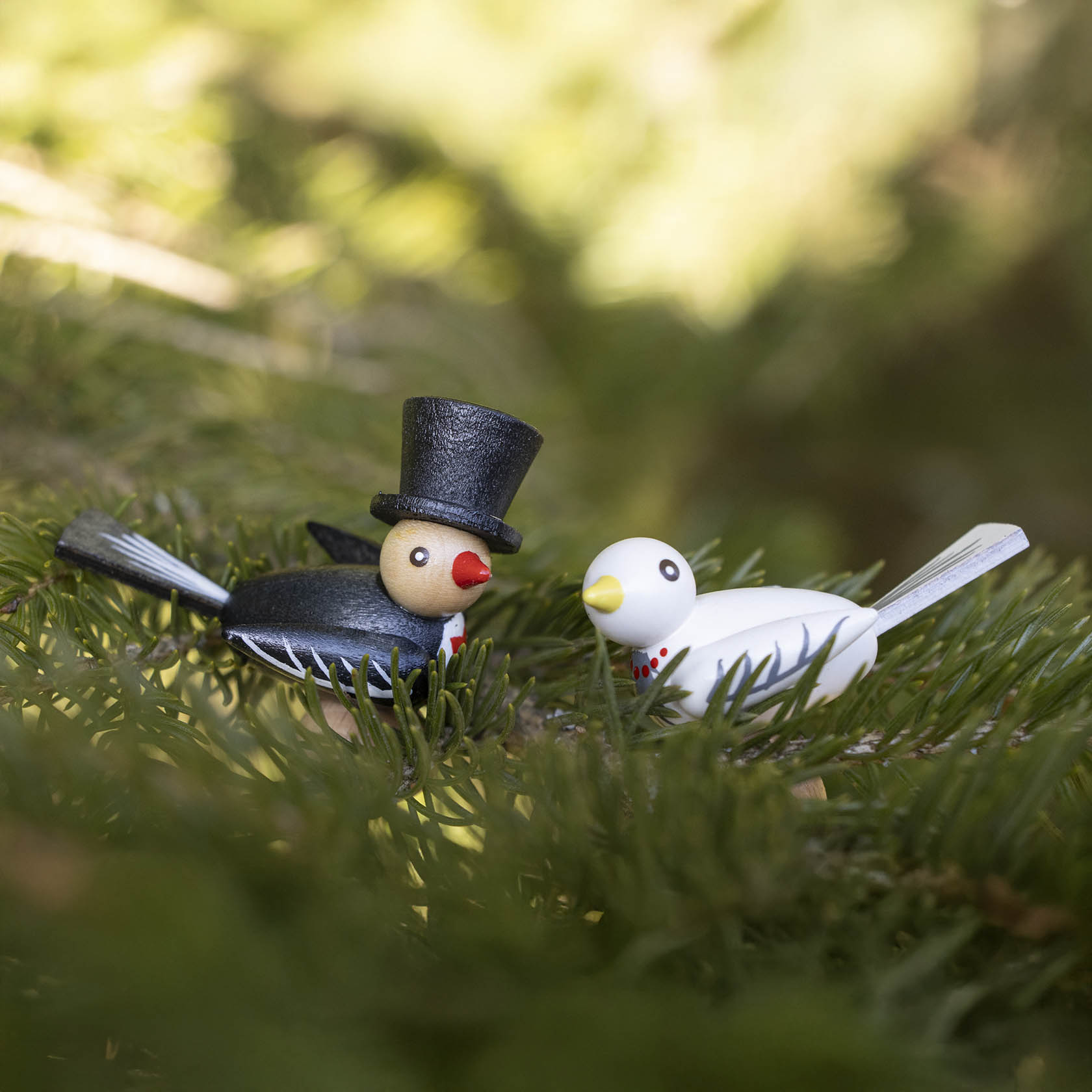 Two bird figurines, one black with a top hat and red beak, and one white with a red beak, sitting on green branches.