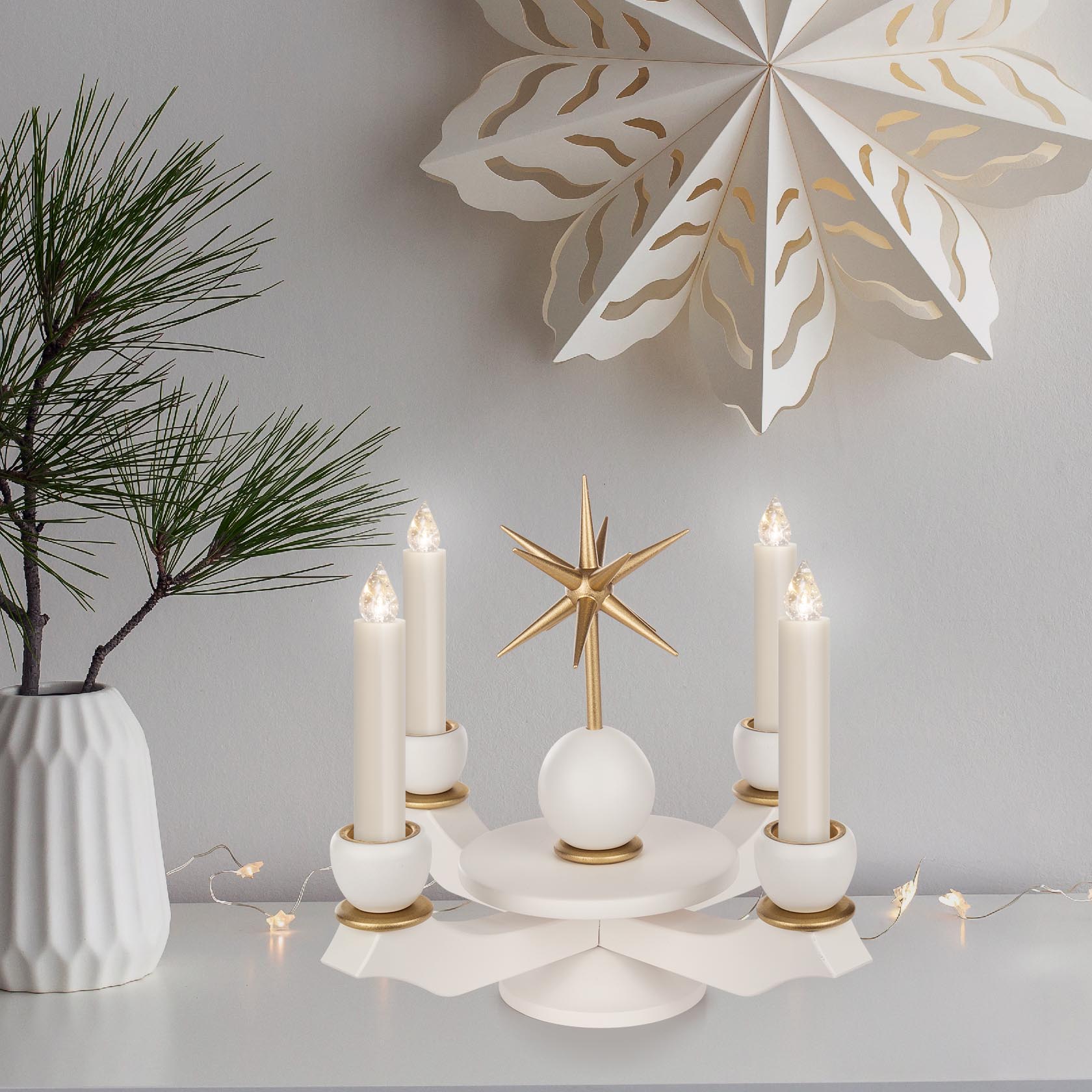 Ein weißer Adventsleuchter mit einem goldenen Stern in der Mitte, umgeben von fünf weißen Wachskerzen auf einem Tisch, geschmückt mit einem Tannenzweig und einer Papierstern-Dekoration im Hintergrund.
