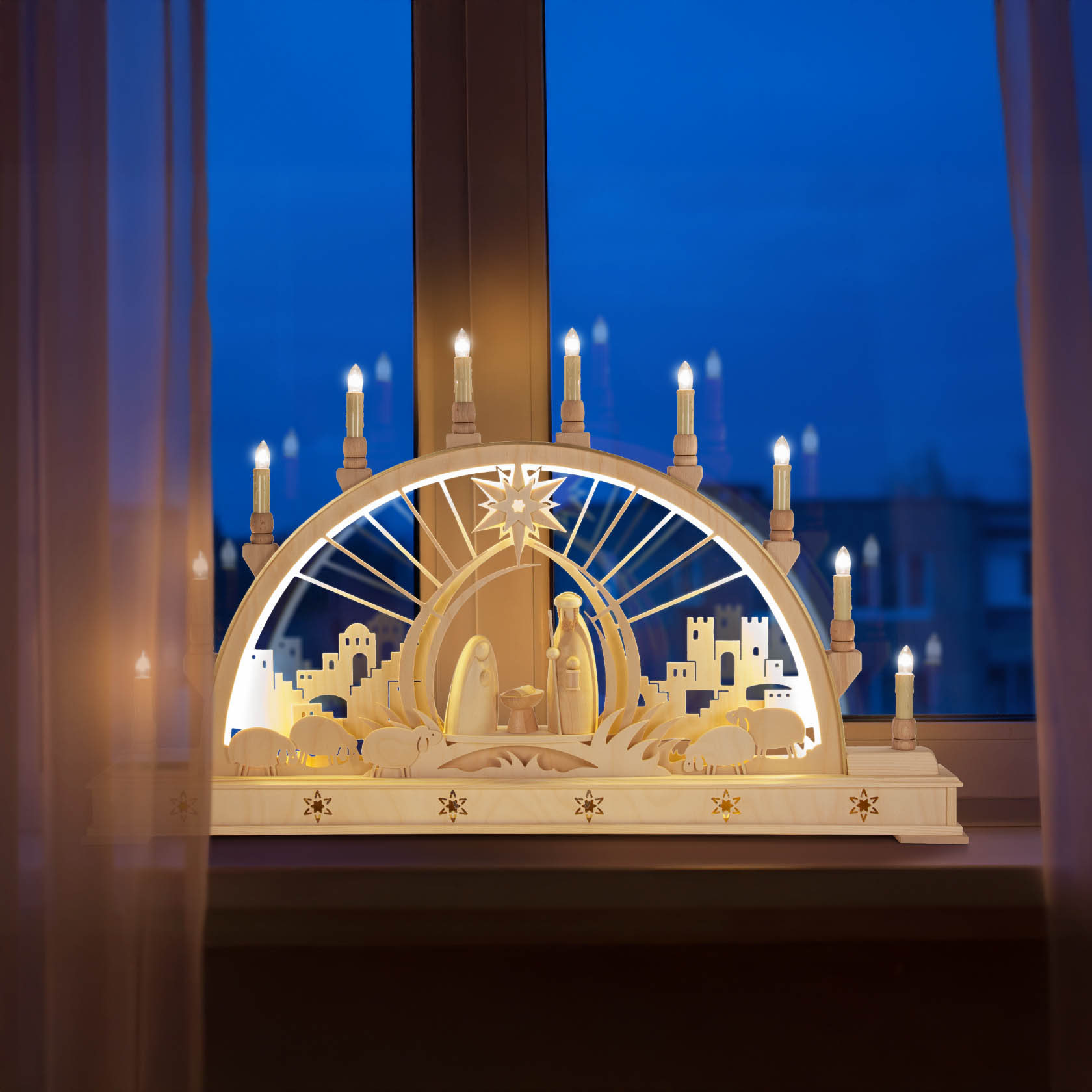 Ein Schwibbogen mit der Darstellung der Geburt Christi, elektrisch beleuchtet, steht auf einer Fensterbank. Die Szenerie zeigt Maria, Josef und das Jesuskind unter einem Sternenhimmel mit Kerzenlichtern.