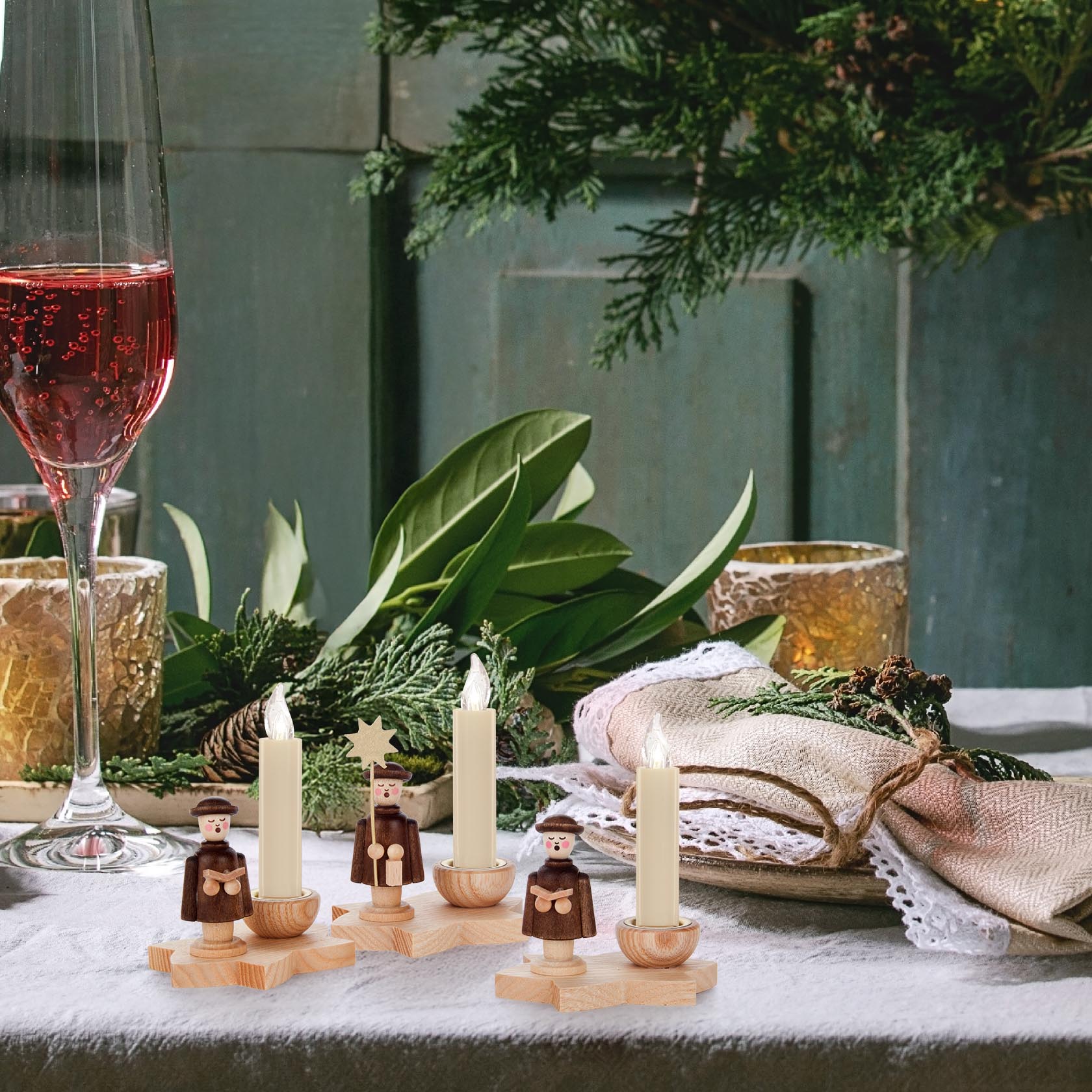 Three wooden candle holders shaped as carol singers, each holding a candle, with a festive table setting including wine and greenery.