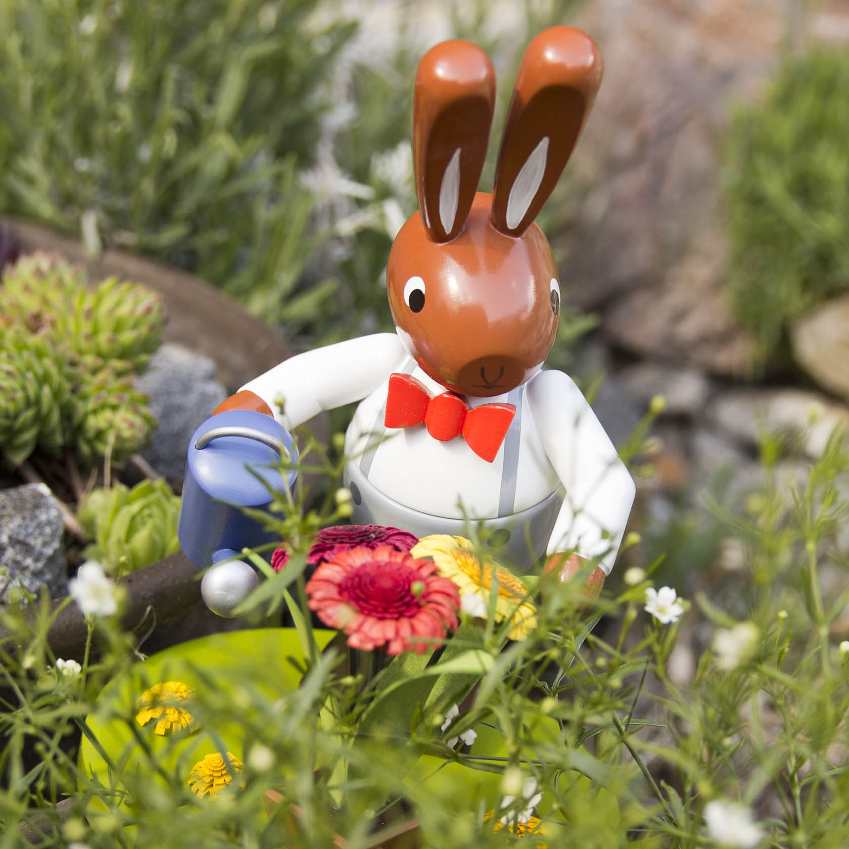 A large plastic rabbit wearing a red bow tie and white shirt, holding a blue watering can, surrounded by colorful flowers.