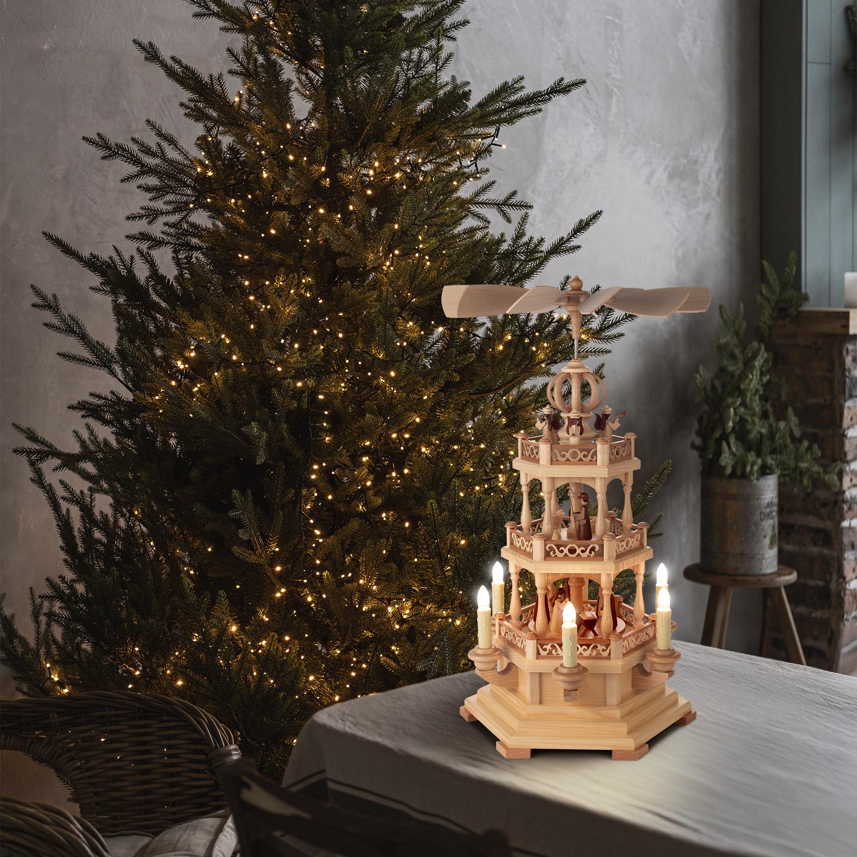 A two-tiered Christmas pyramid with a nativity scene, featuring electric lighting and movement, set on a table next to a lit Christmas tree.