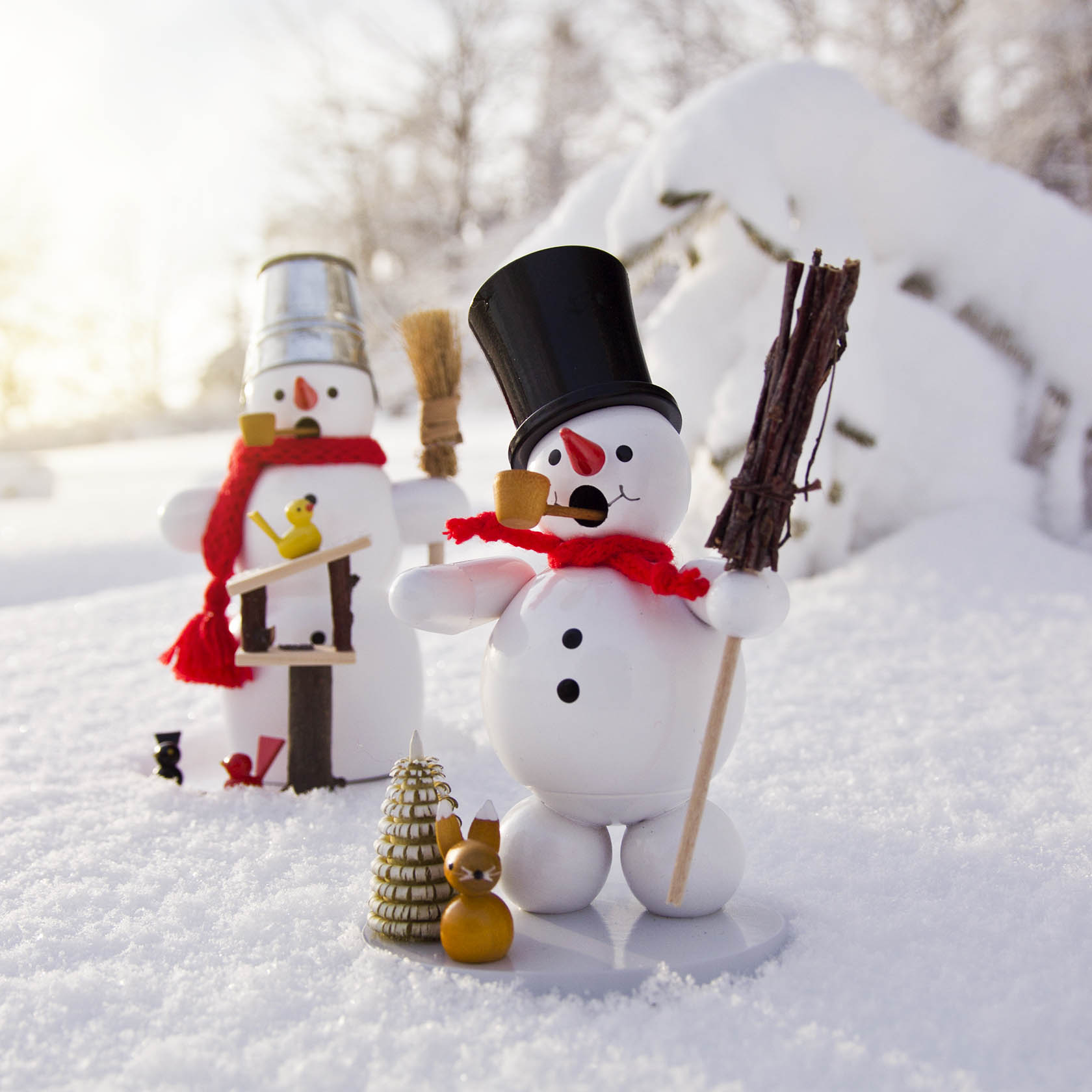 Vordergrund: Schneemann-Räuchermann mit schwarzem Zylinder, Pfeife und rotem Schal in verschneiter Landschaft. Neben ihm ein Mini-Bienenstock und ein Eichhörnchenfigur. Hintergrund: Zweiter Schneemann mit Metalleimer auf dem Kopf, roter Schal, hält Vogelh