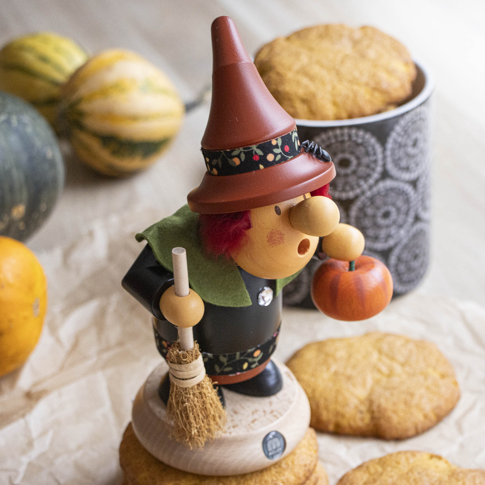 A carved wooden smoking figure of a Halloween witch with a pointed hat, holding a small broom and pumpkin, standing on cookies next to a patterned cup and gourds.