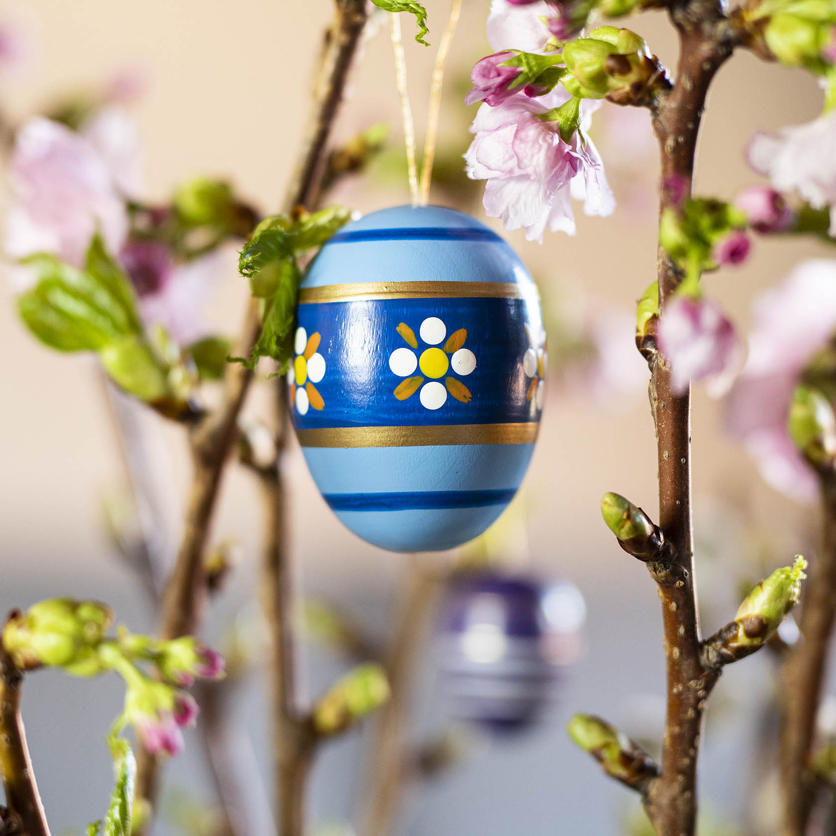 Ein blaues Osterei mit goldenen Streifen und weißen Blumenmustern hängt an einem Zweig mit rosa Blüten.