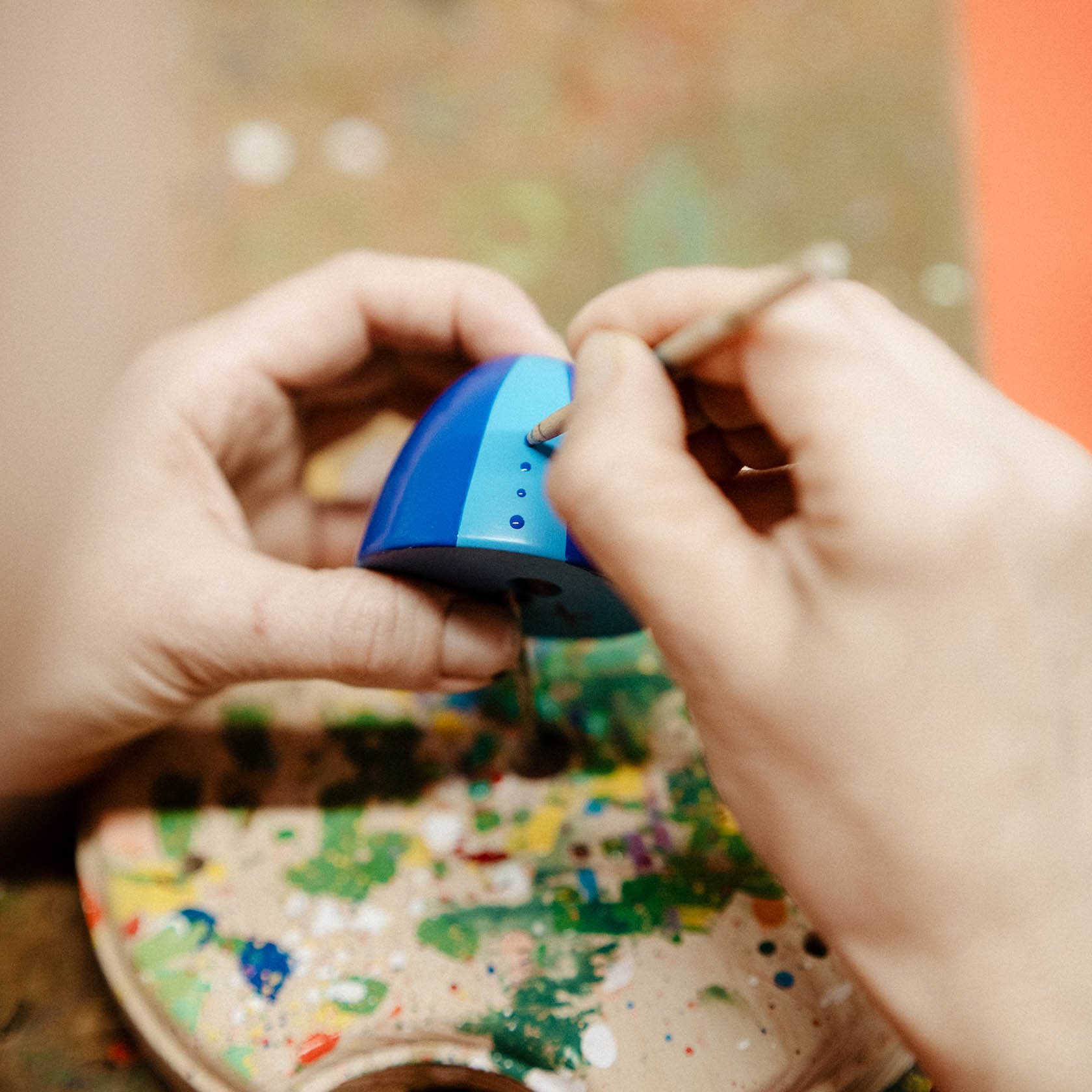 Hands of a person meticulously painting small dots on a blue figurine with a thin brush.