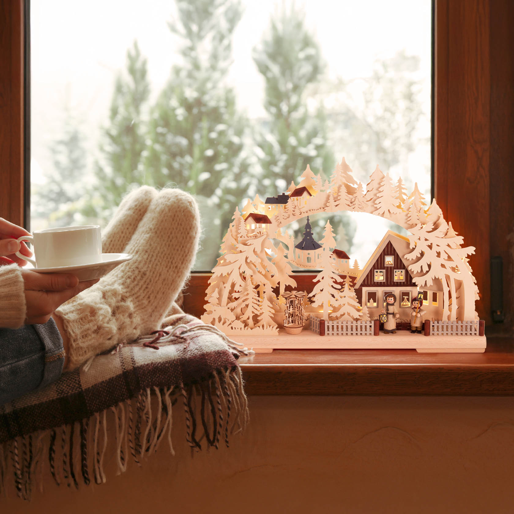 A cozy scene showing a Schwibbogen with electric lights depicting a village with houses and trees on a windowsill. A person with knitted socks holds a cup.