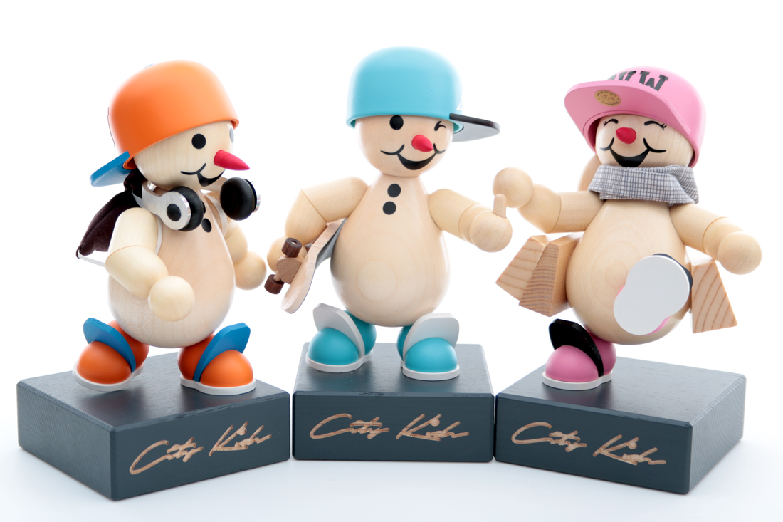 Drei Holzpuppen in modischer Kleidung: eine mit orangefarbener Mütze und Kopfhörern, eine mit blauer Mütze und Skateboard und eine mit pinker Mütze und Schal.