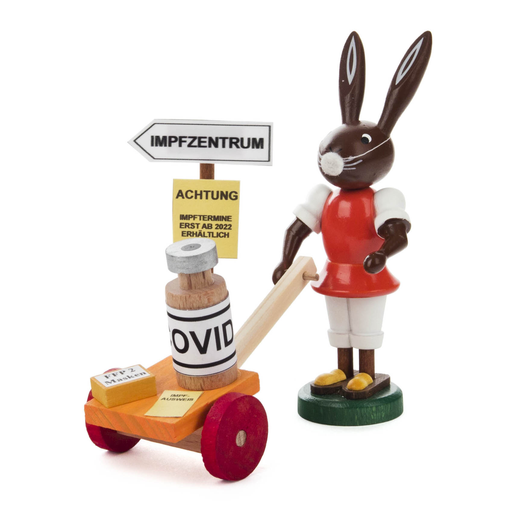 Eine hölzerne Hasenfigur in Rot-Weiß zieht einen Wagen mit einem überdimensionalen Impfstofffläschchen. Schilder darauf verweisen auf Covid-Impfungen. im Dregeno Online Shop günstig kaufen
