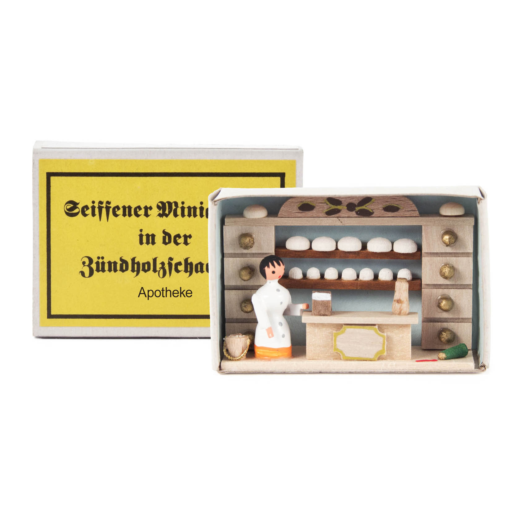 A miniature pharmacy scene inside a matchbox showing a female pharmacist in a white coat, standing behind a counter with shelves filled with small medicine jars. im Dregeno Online Shop günstig kaufen