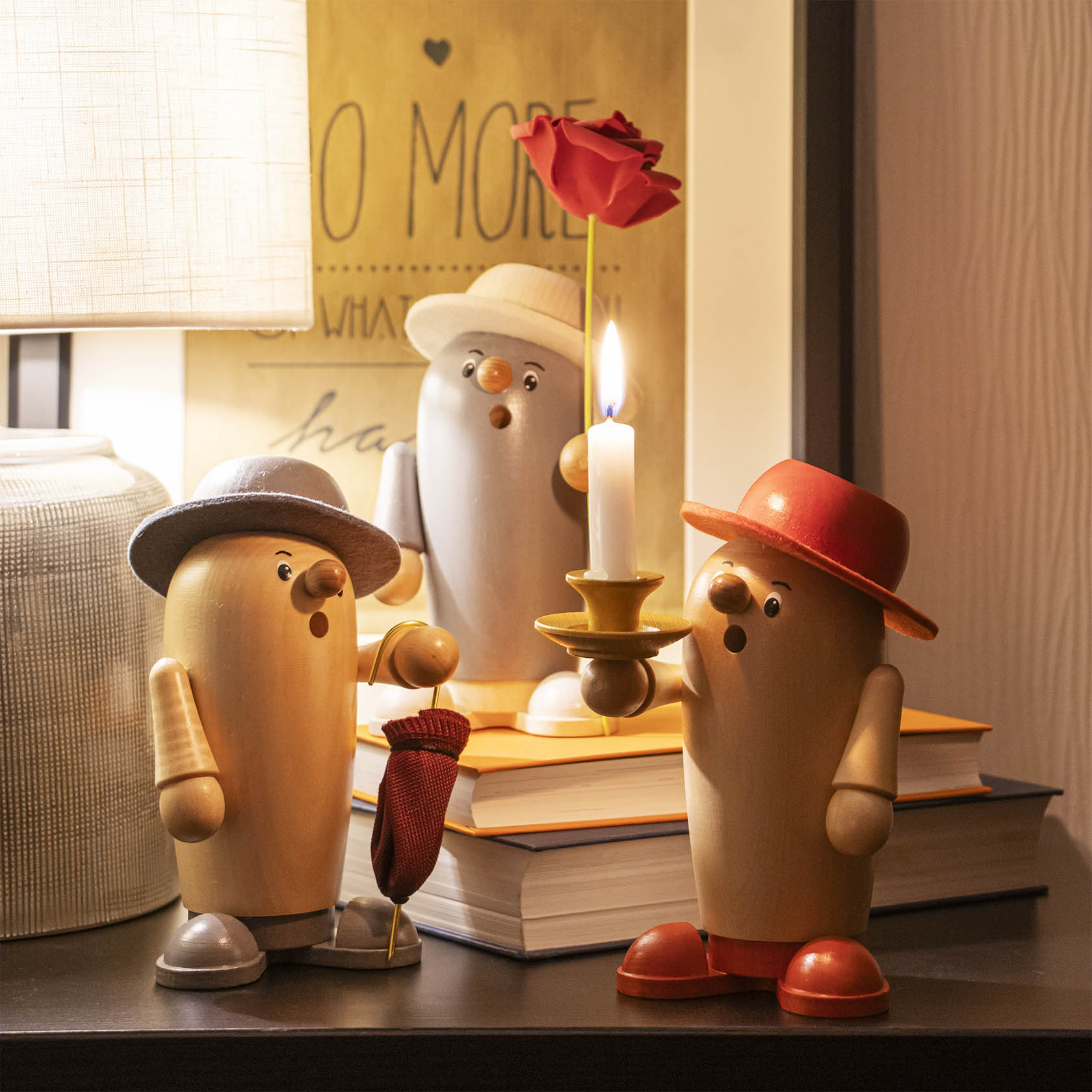 Three wooden smoking figures on a shelf: one in grey/white with a rose and grey hat, another holding a candle with a red hat, and a third with a grey hat holding a pipe.