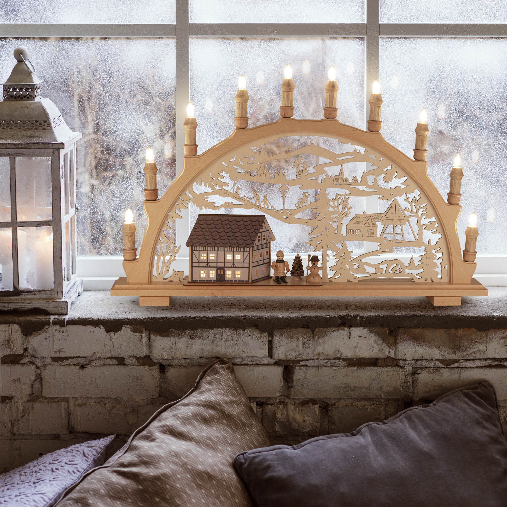 Ein filigraner Schwibbogen mit LED-Beleuchtung zeigt Winterlandschaft mit Fachwerkhaus und Winterkindern, platziert auf einer Fensterbank.