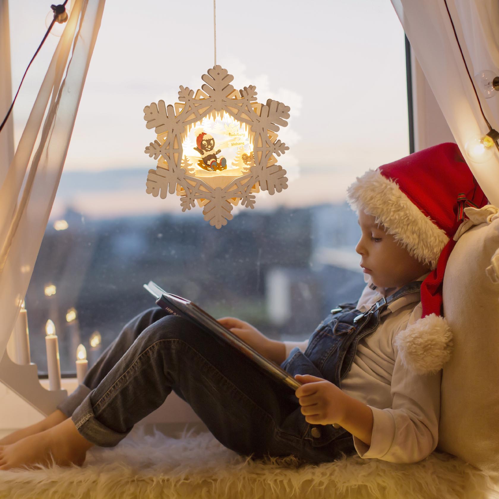 Young child in Santa hat sitting by window, reading a book with an illuminated snowflake decoration hanging above.