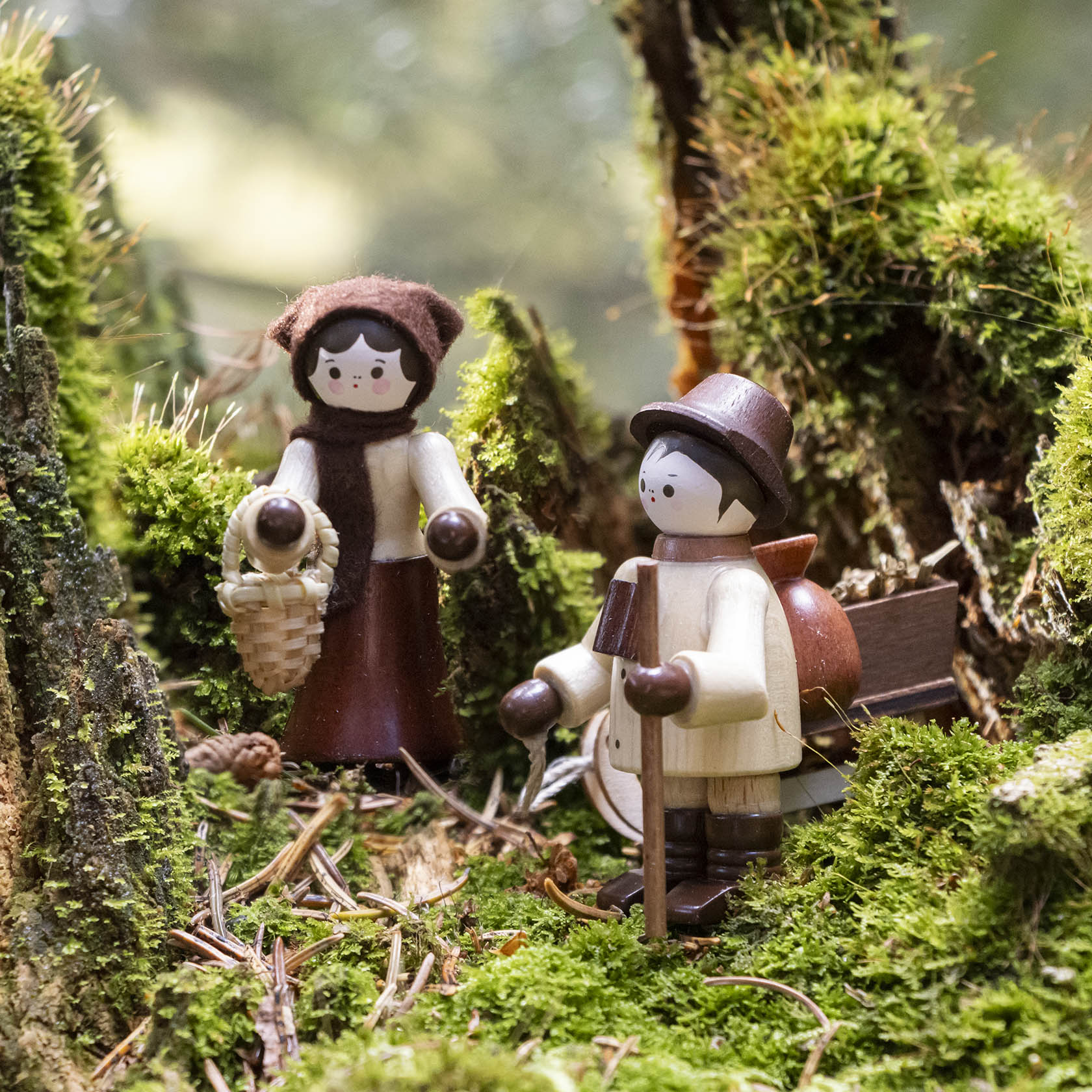 Zwei Holzfiguren im Wald: Eine Frau mit Kopftuch und Korb und ein Mann mit Hut und Wanderstock stehen zwischen Moos und Holz.