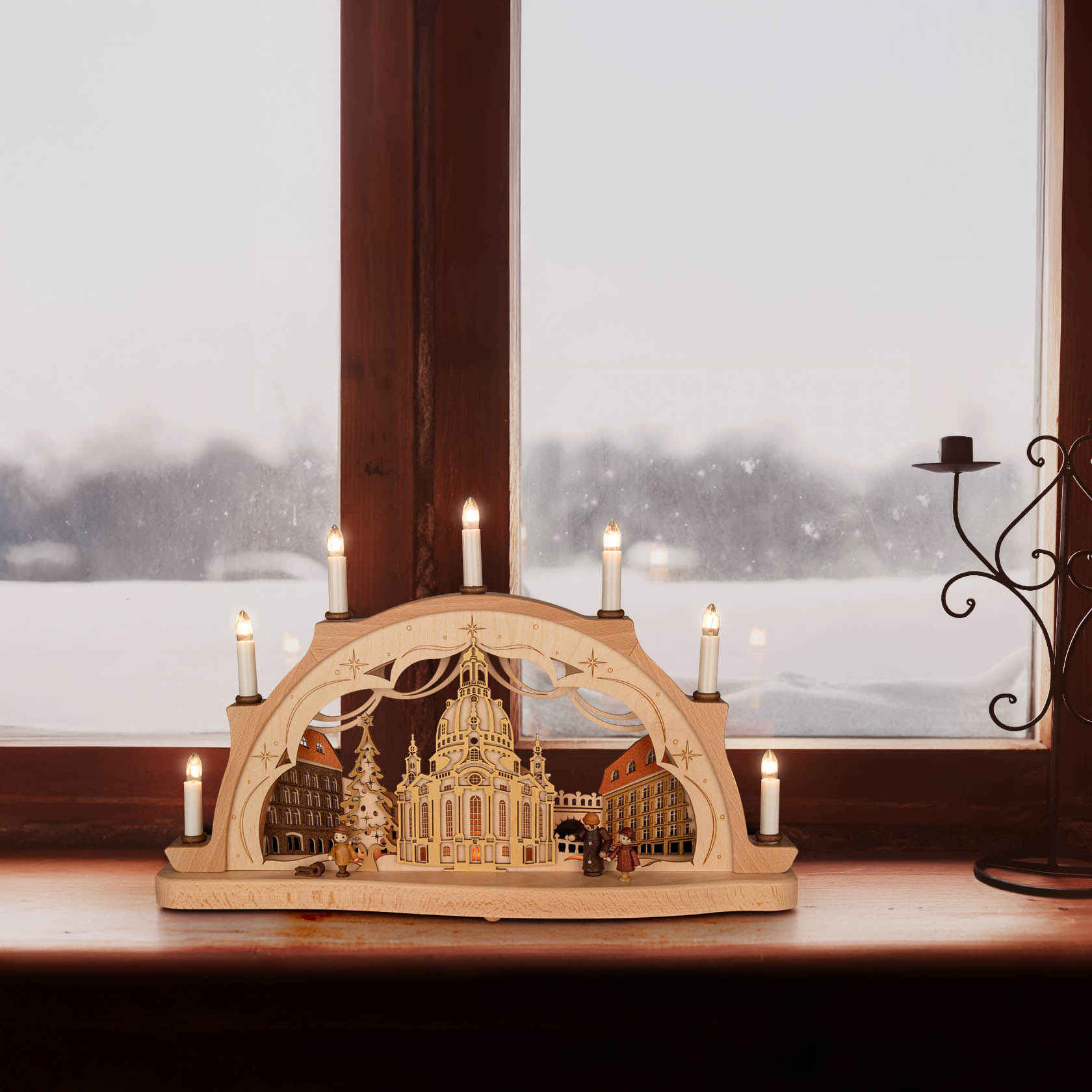 A 3D Schwibbogen displaying Dresden's Frauenkirche with electric lighting, placed on a windowsill, framed by a snowy winter scene outside.