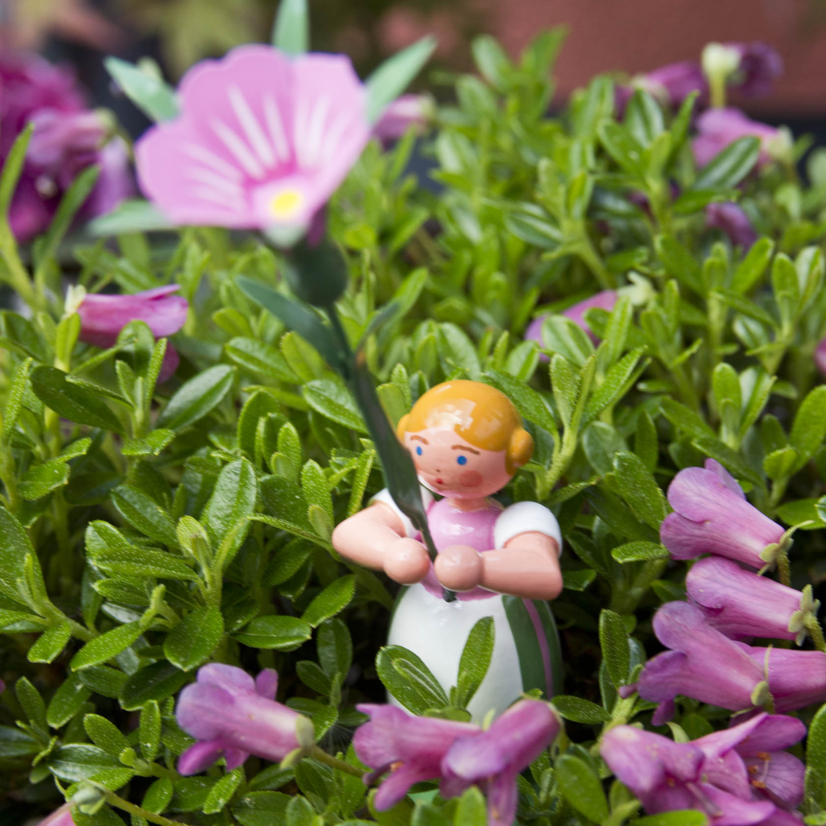 A toy figure of a girl with orange hair holding a pink flower, surrounded by green foliage and small pink blossoms.