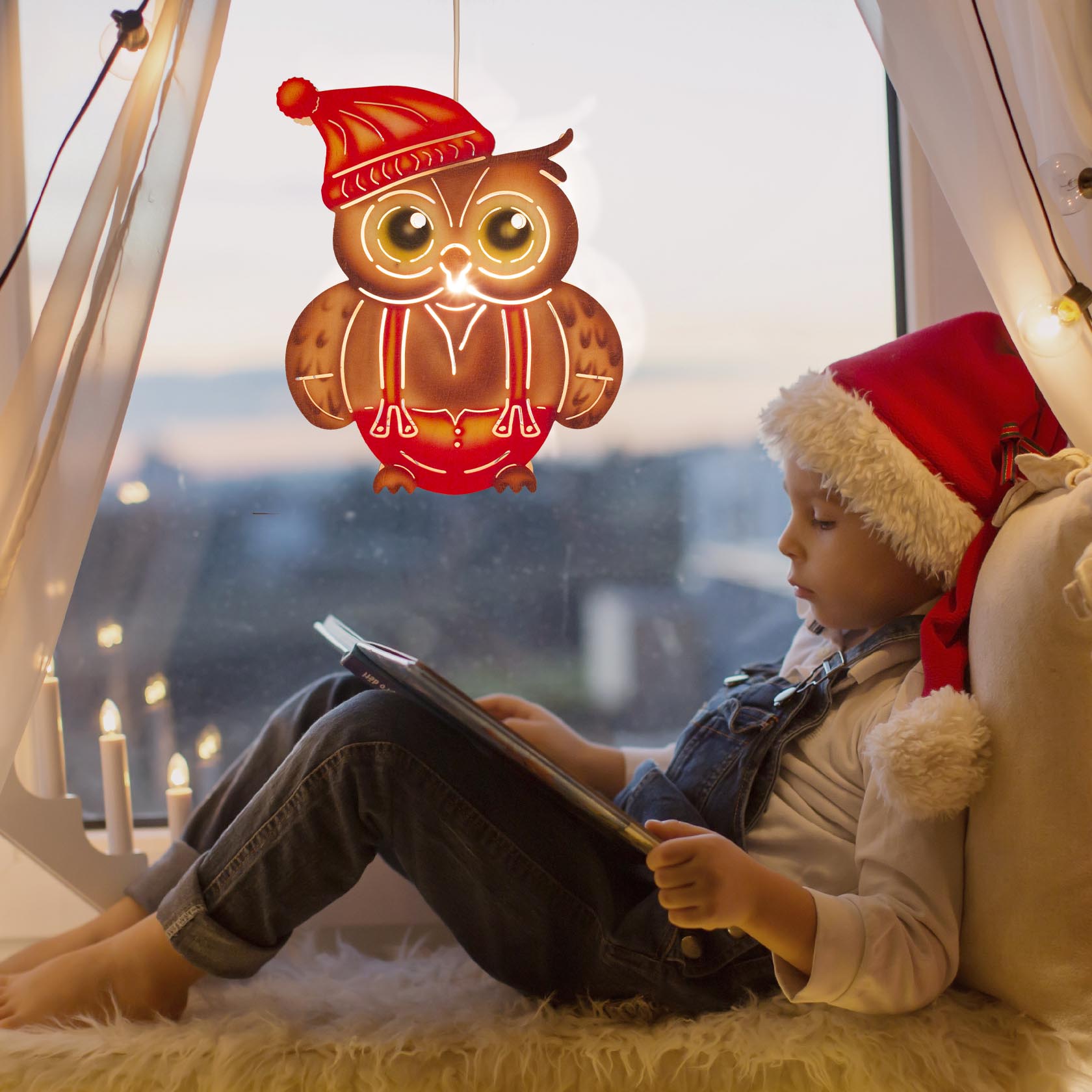 Ein Junge mit Weihnachtsmütze sitzt bei Kerzenlicht am Fenster und liest, während eine leuchtende Eule mit roter Mütze und elektrischer Beleuchtung am Fenster hängt.