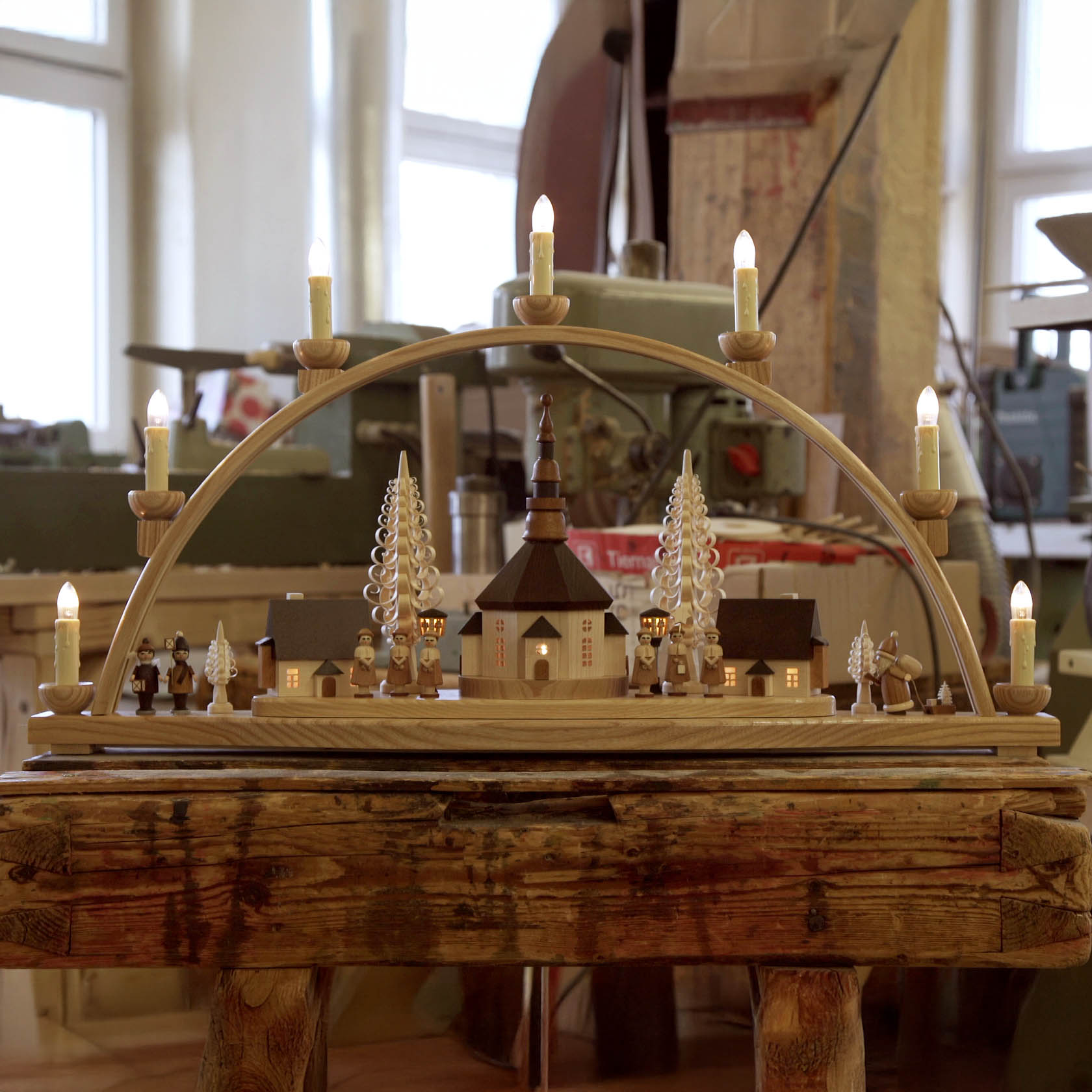 Illuminated wooden arch featuring a Seiffener village scene with a church, lit buildings, lanterns, and figurines arranged on a rustic table.