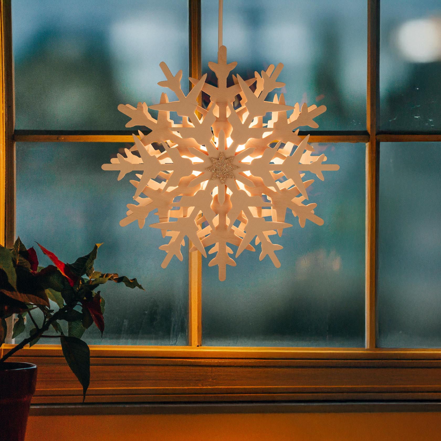Ein beleuchteter Schneekristall hängt vor einem Fenster. Der Kristall hat eine elektrische Beleuchtung und strahlt warmes Licht aus.