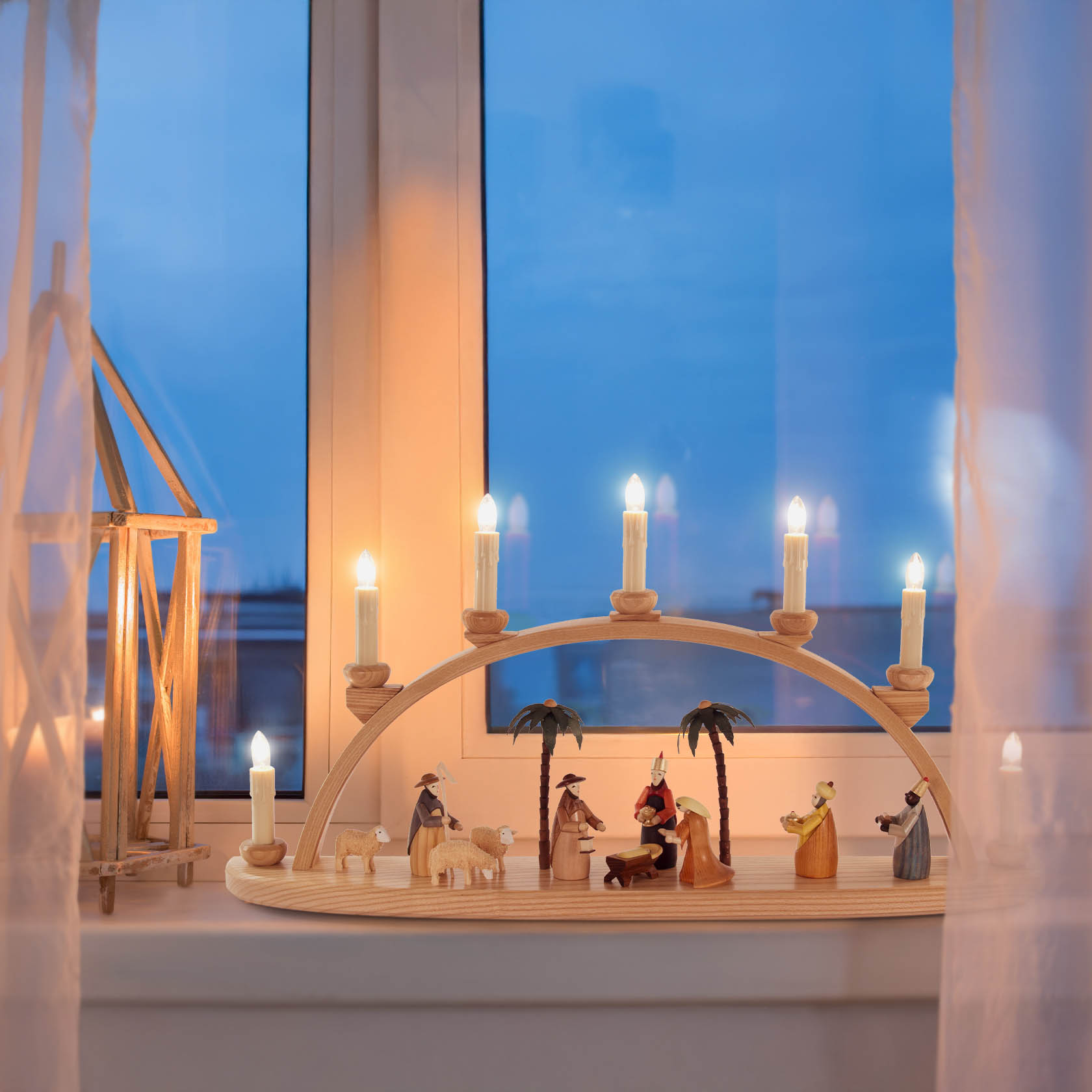 Ein farbiger Schwibbogen mit der Darstellung der Geburt Christi. Elektrisch beleuchtet mit Kerzenlichtern, auf einem Fensterbrett bei Dämmerung platziert.