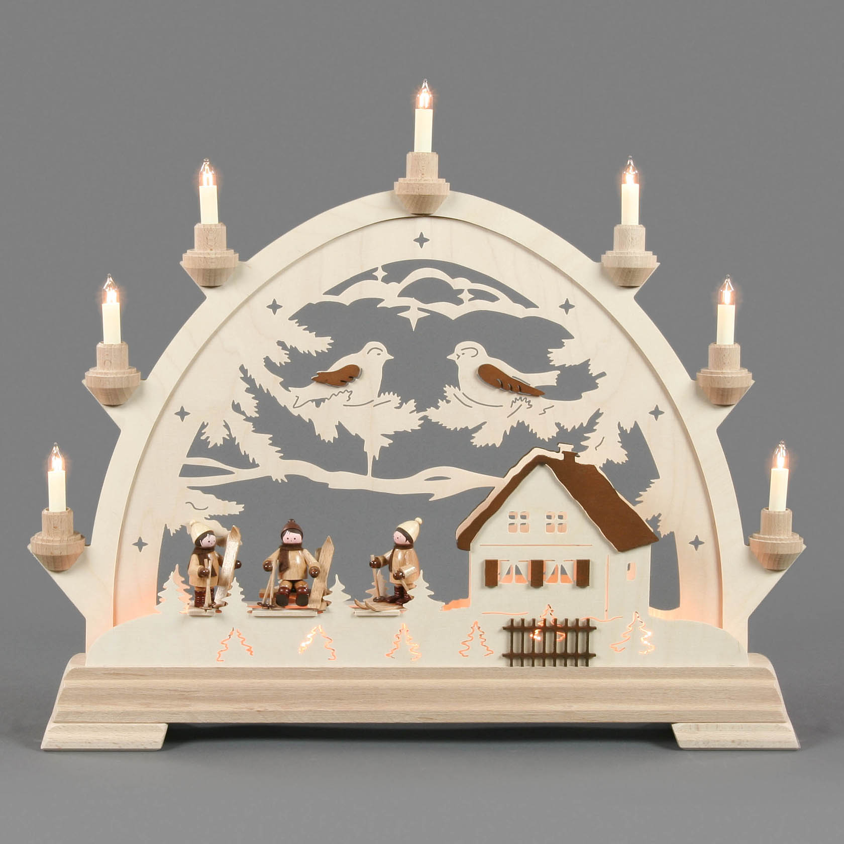 An electric candle arch with skiers, featuring a cozy house and two birds on branches, all illuminated by small lights.