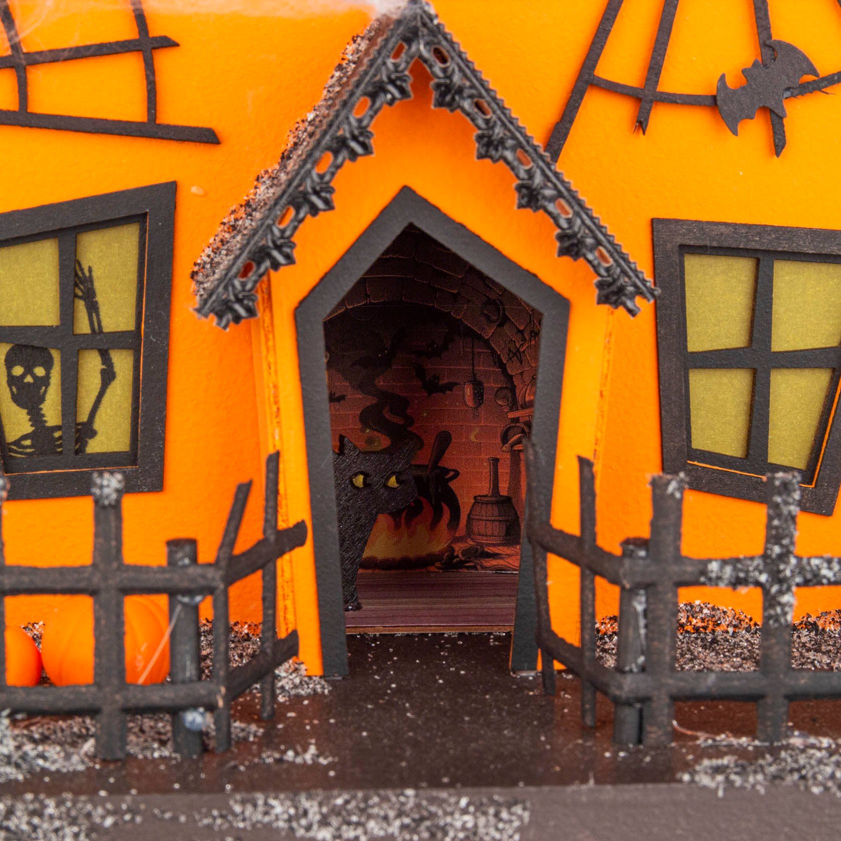 Ein dekoratives Kürbishaus mit schiefen schwarzen Holzfenstern und Türrahmen. Eine Skelettfigur sowie ein Hexenkessel sind im Inneren sichtbar.