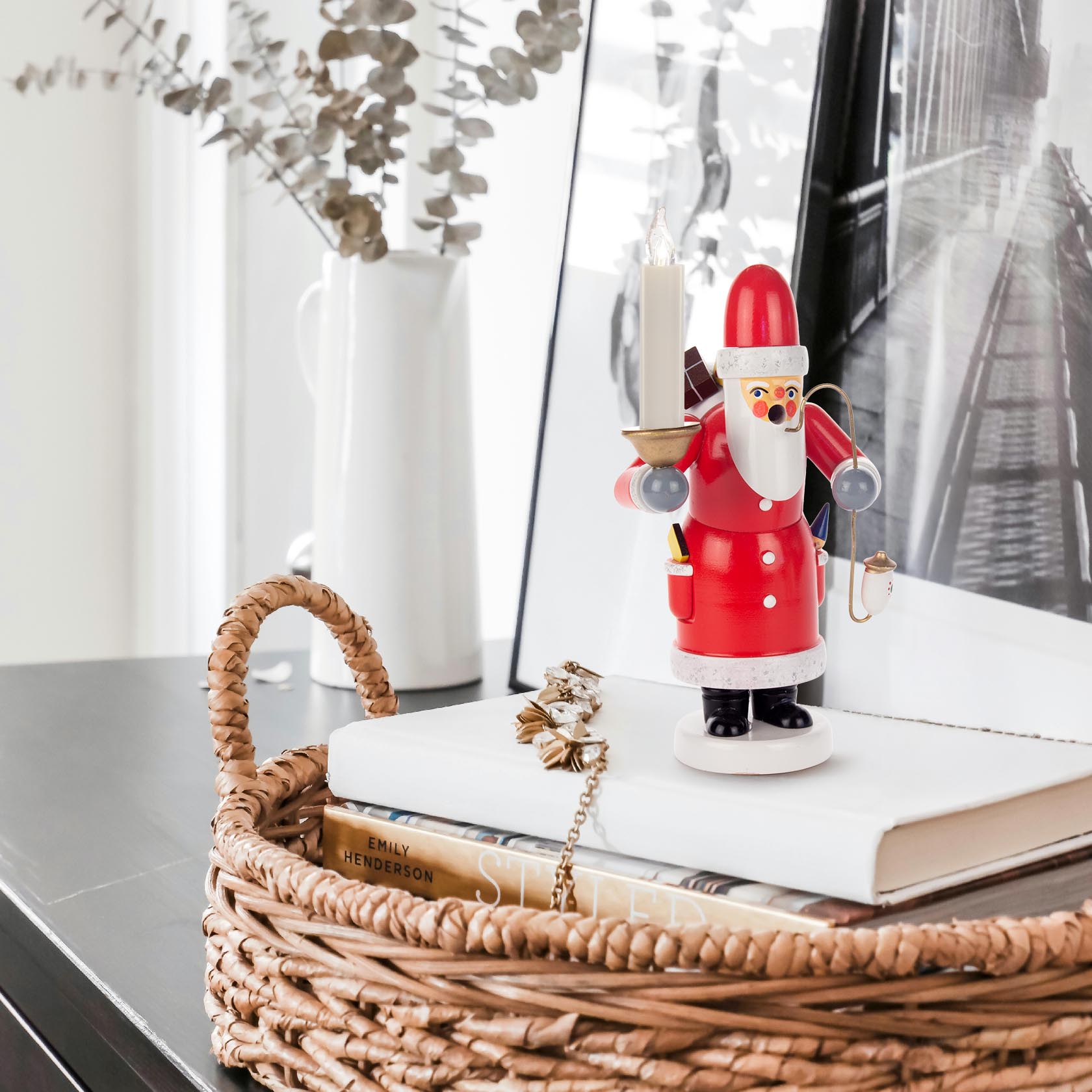 Ein traditioneller Räuchermann in Form eines Weihnachtsmanns, der einen Kerzenhalter mit einer angezündeten Kerze trägt. Er steht auf einem Tisch neben einem Weidenkorb.