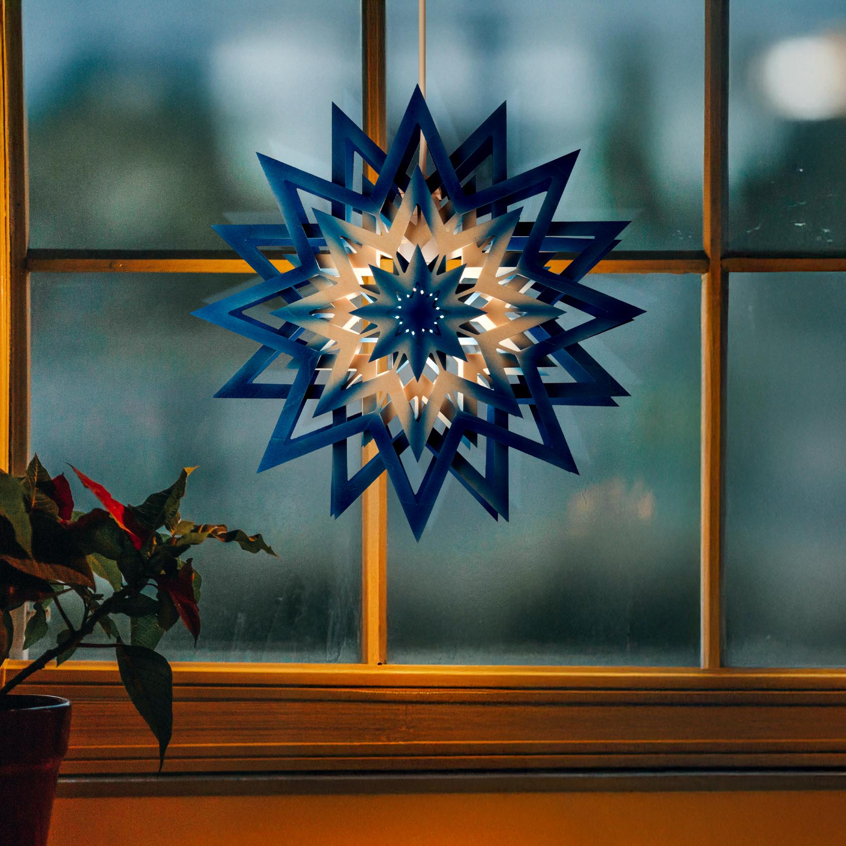 Blue and natural-colored star-shaped light decoration hanging in a window, illuminated with electric lighting.