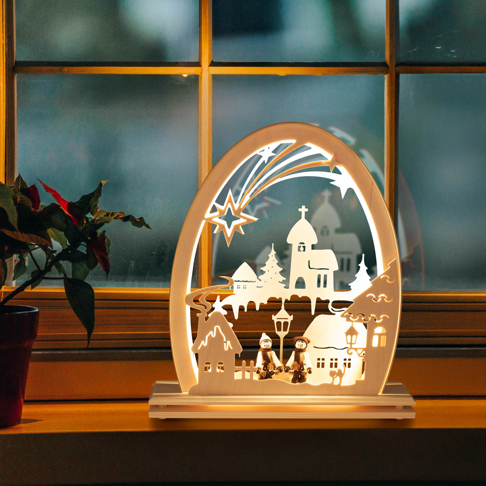 An illuminated LED arch depicting a winter village scene with a church, trees, houses, and two figures sitting on a bench under a streetlamp.