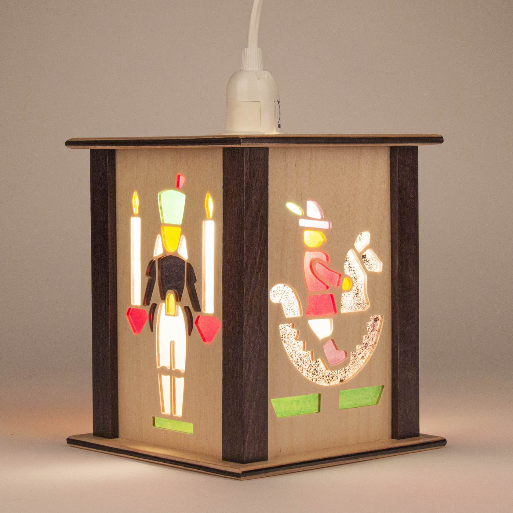 Wooden lantern featuring cut-outs of a soldier with candles and a boy on a rocking horse, both illuminated from within.