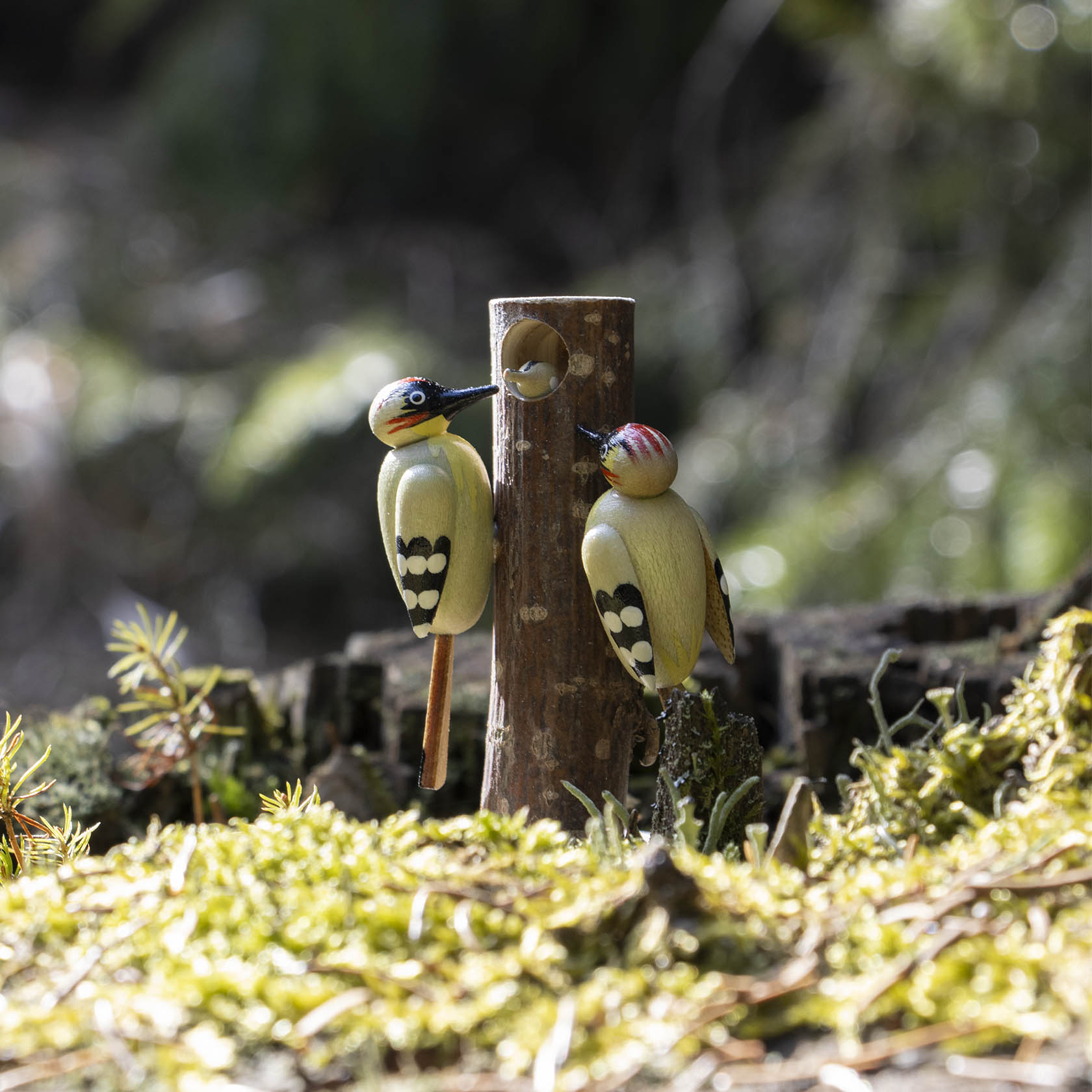 Two wooden green woodpeckers, one adult and one juvenile, are perched on a wooden post with a small hole, depicting a family of birds.