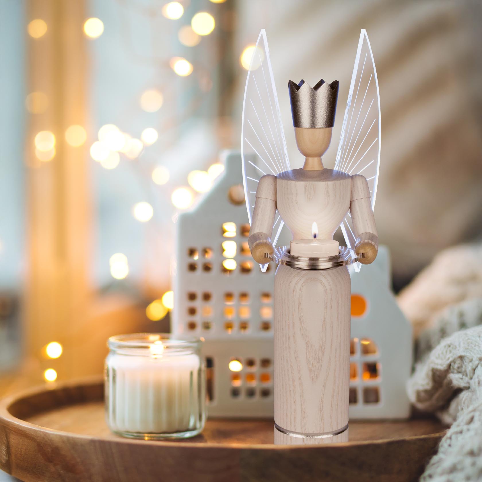 A wooden angel figure with LED-lit wings, wearing a crown, standing on a wooden tray next to a lit candle in a cozy setting.