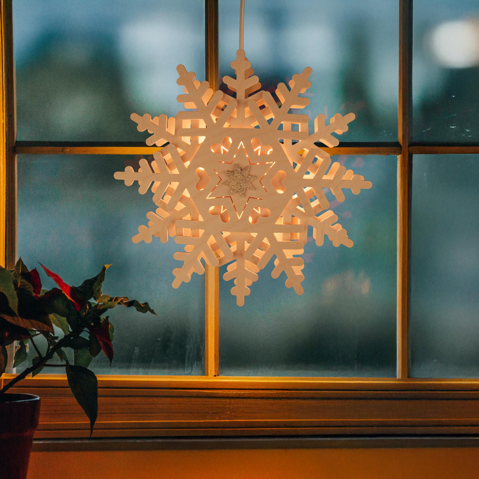 Fensterbild eines beleuchteten Schneekristalls vor einem Fenster. Der Schneekristall ist elektrisch erleuchtet und verbreitet warmes Licht.