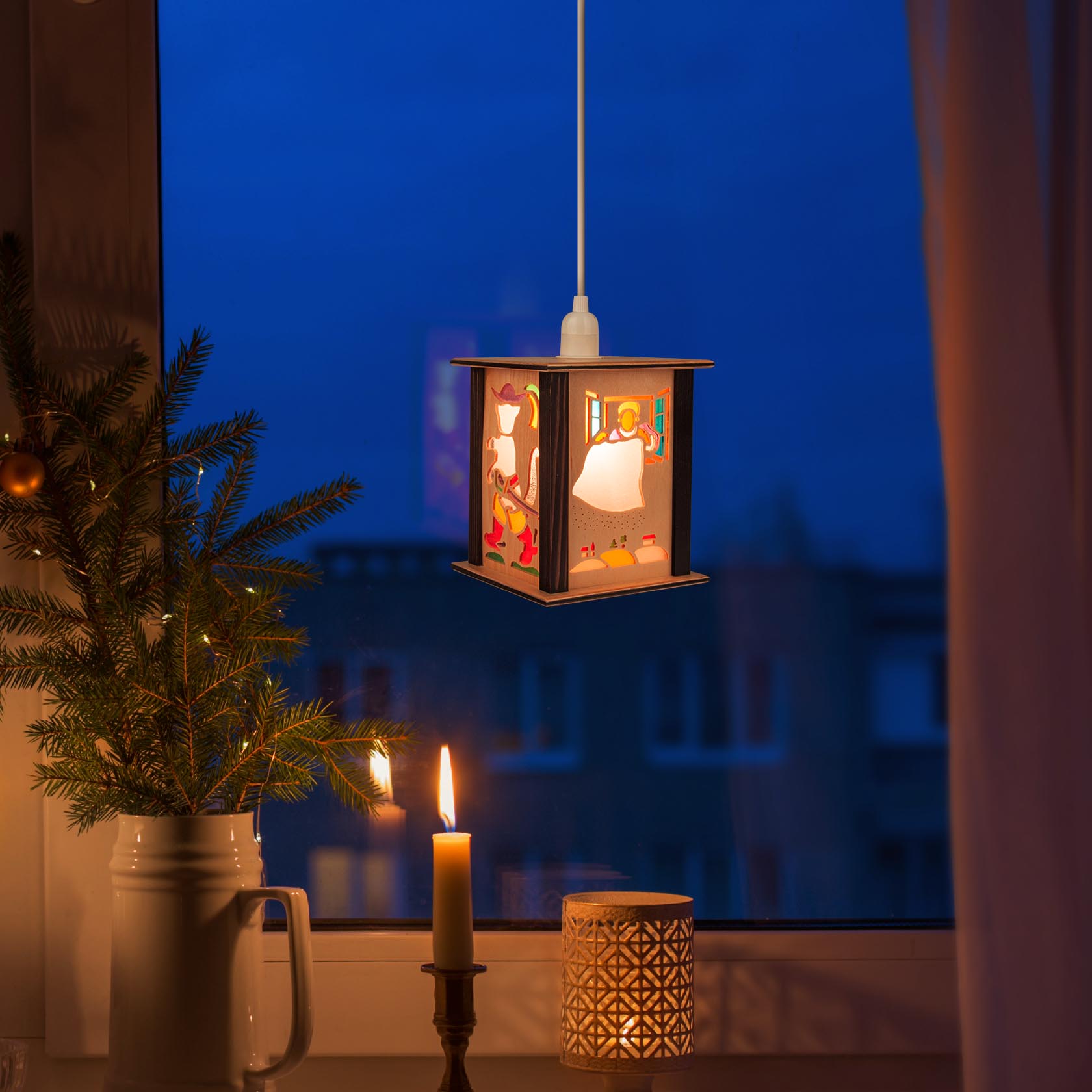 A small lantern with fairy tale illustrations, hanging on a window at dusk. A lit candle and a small decorated tree are on the windowsill.
