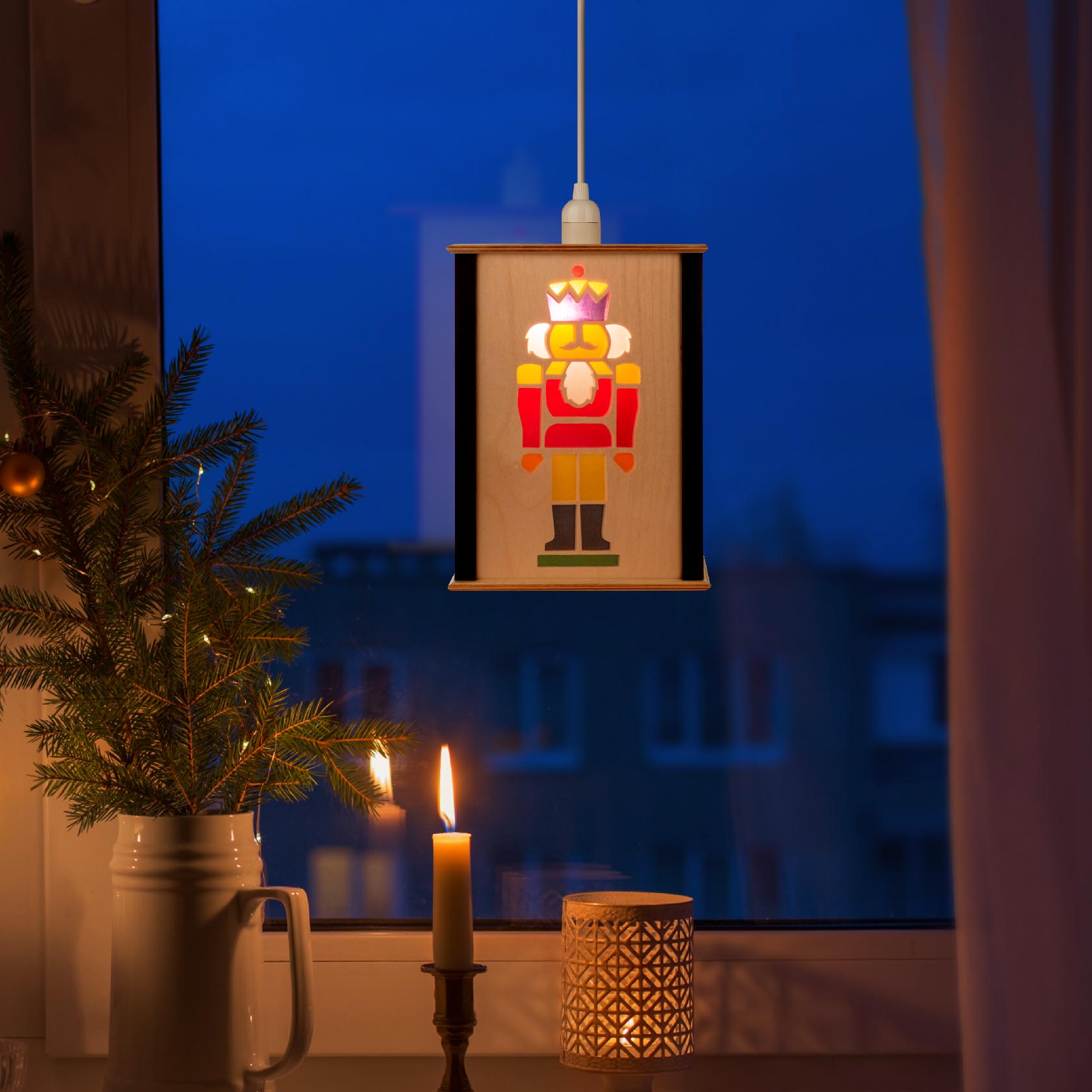 A small window lantern featuring a colorful nutcracker design, glowing warmly in the evening. It hangs from the top with a visible light bulb.