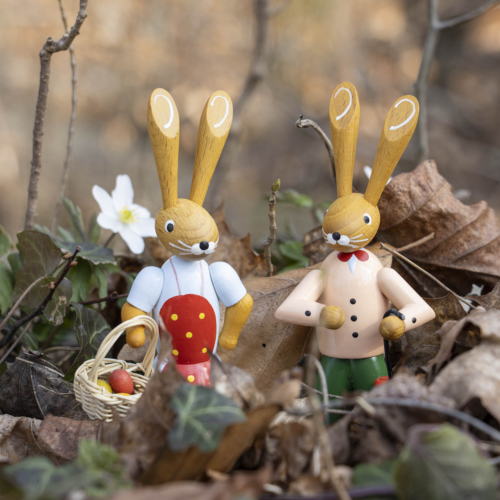 Two wooden bunny figurines standing side by side; one wears a red apron with yellow dots and holds a basket, and the other wears a beige coat, surrounded by leaves.