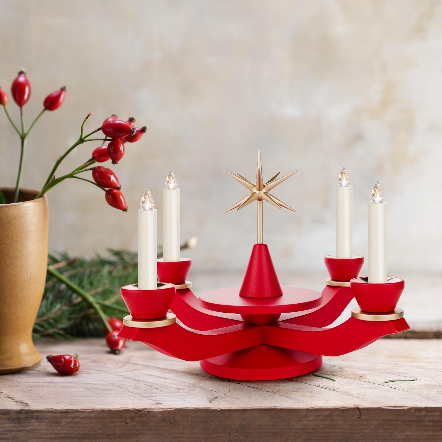 Roter Adventsleuchter mit vier weißen Kerzen und einem goldenen Stern in der Mitte, auf einem Holztisch dekoriert mit Hagebuttenzweig und Tannenzweig.