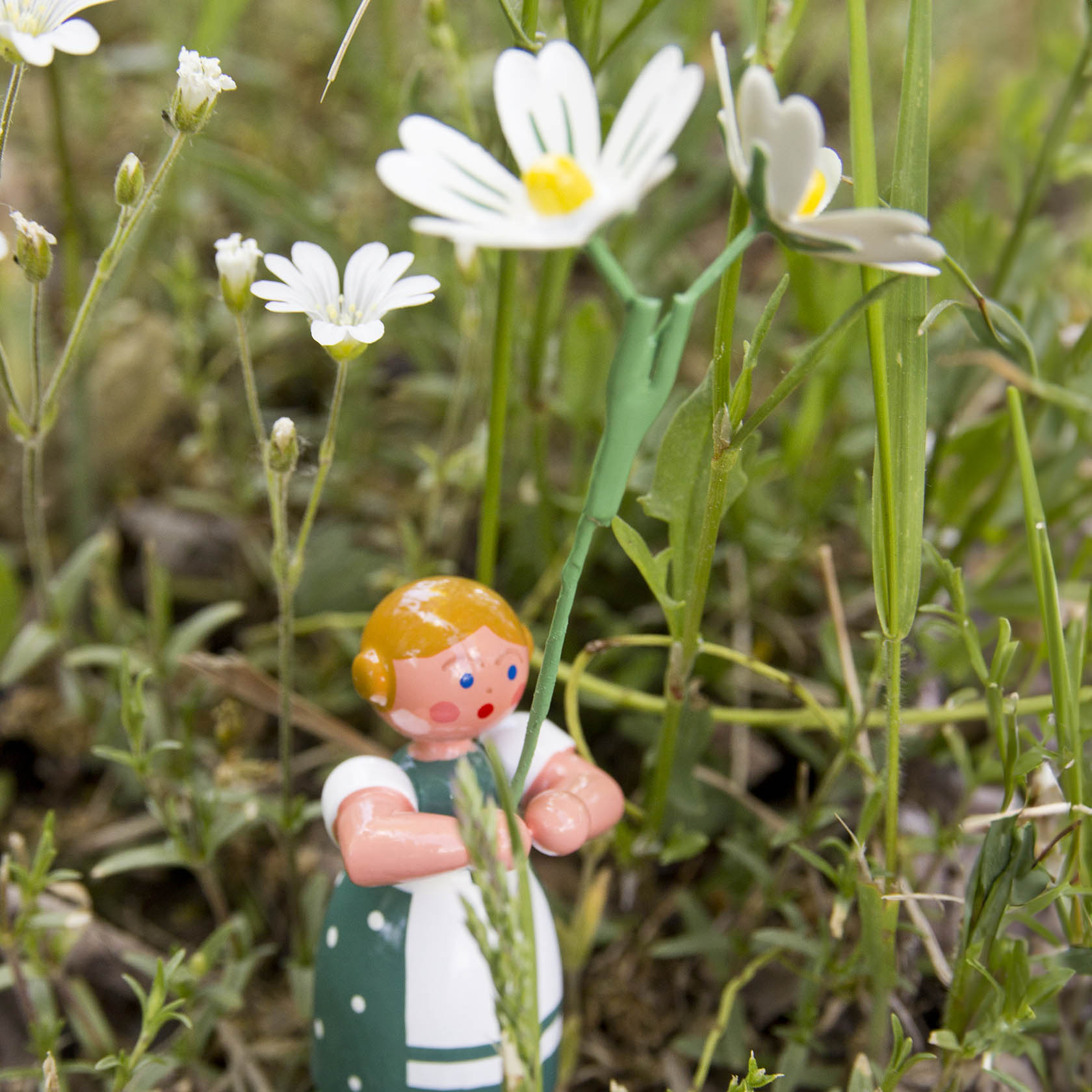 Holzfigur eines Mädchens mit orangefarbenen Haaren im grünen Kleid, hält eine weiße Sternmiere in der Hand. Die Figur steht inmitten von Gras und weiteren Sternmieren.