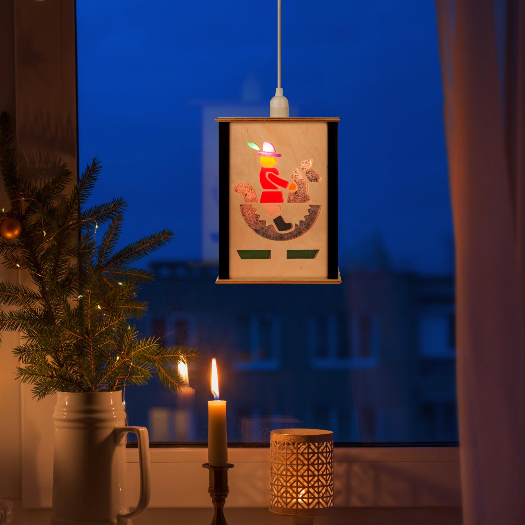 A small lantern hanging in a window, featuring an illustration of a rider on a rocking horse, illuminated from inside. A lit candle and a small pine decoration are nearby.