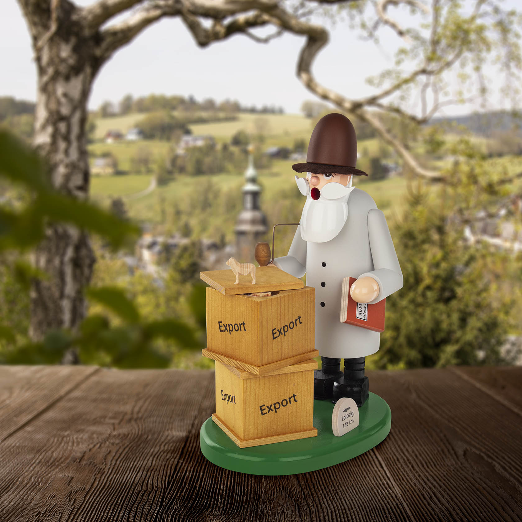 Wooden smoker figure resembling a publisher, holding a book and standing next to two labeled export boxes, set against a scenic countryside.