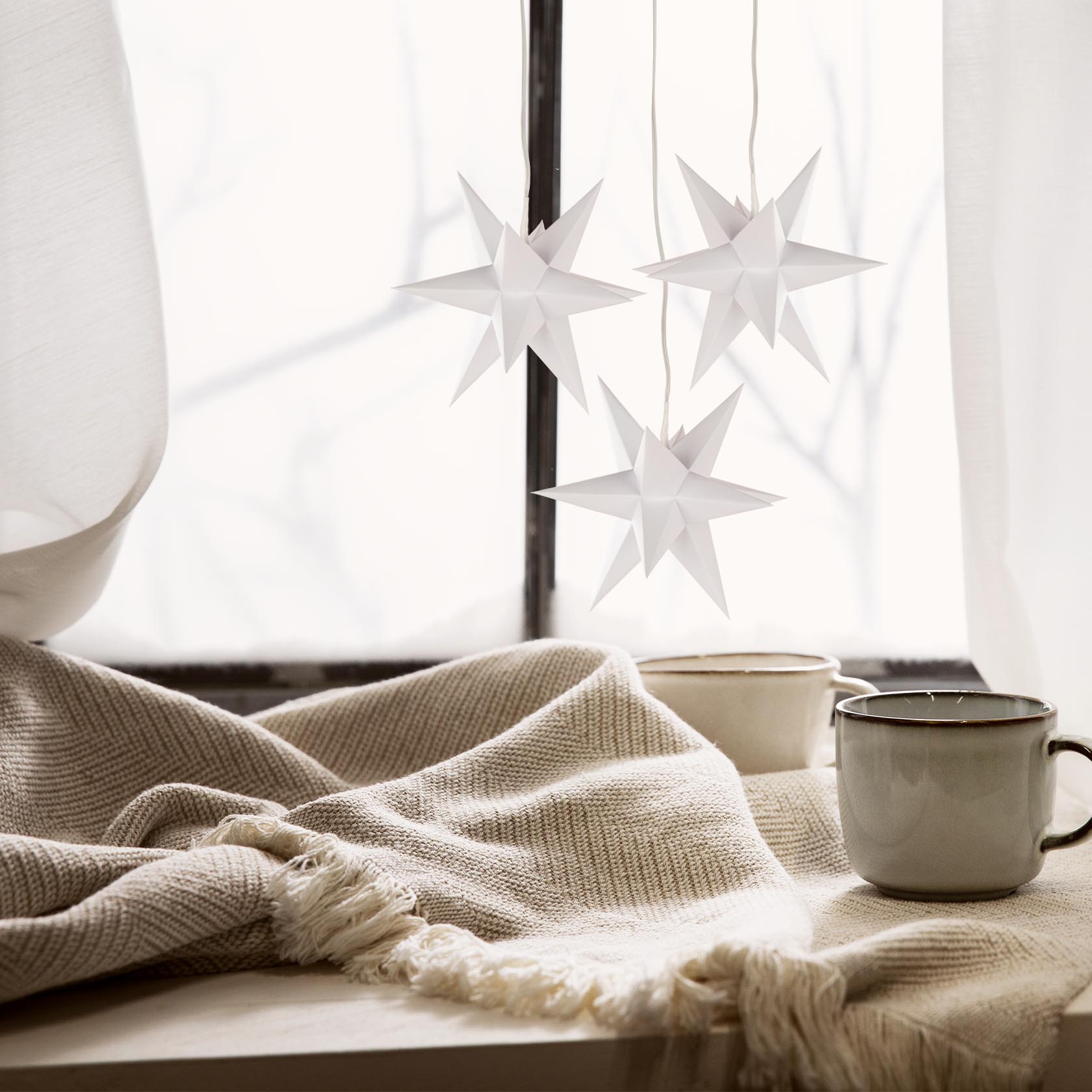 Drei weiße, einfarbige Mini-Adventssterne mit elektrischer Beleuchtung hängen vor einem Fenster. Daneben stehen zwei Tassen auf einem mit Decke bedeckten Tisch.