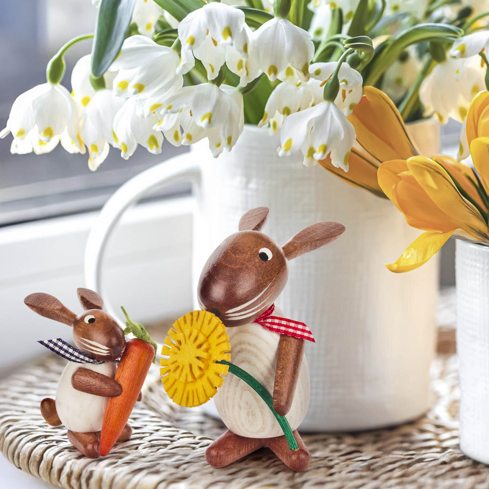Two wooden rabbit figurines; one holds a spring flower, the other a carrot. Both are white and brown with checkered scarves.
