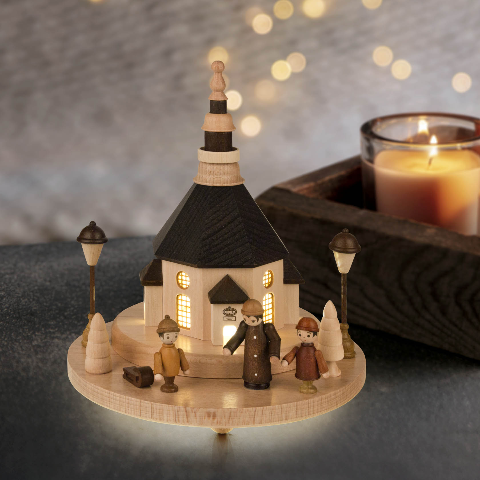 A wooden model of the Seiffener Church with market figures illuminated by LED lights. The scene includes three small wooden figures, two lamp posts, and trees.