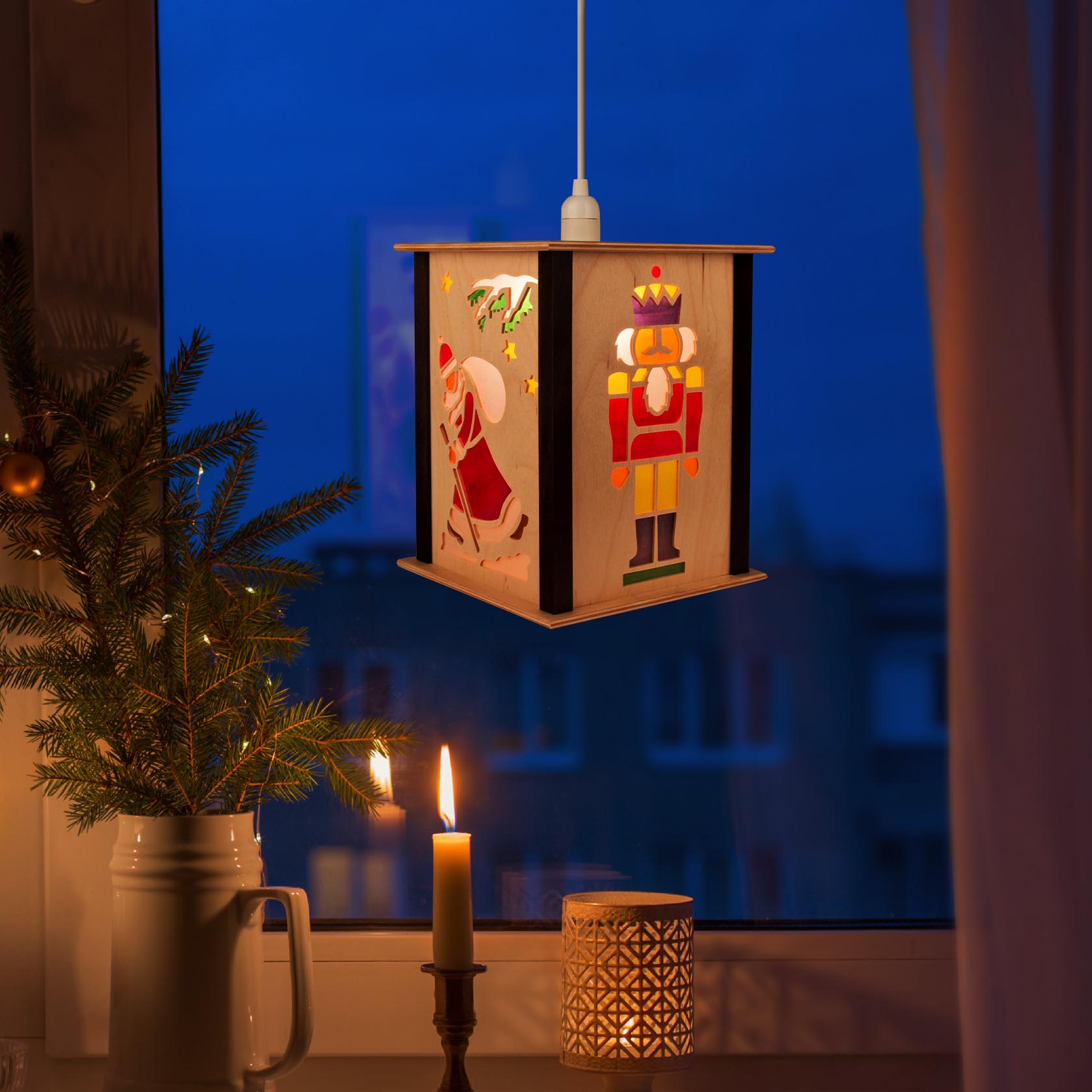 Eine hängende Laterne mit Nussknacker-Motiv, daneben ein leuchtendes Teelicht und ein kleiner Tannenzweig in einer Vase.