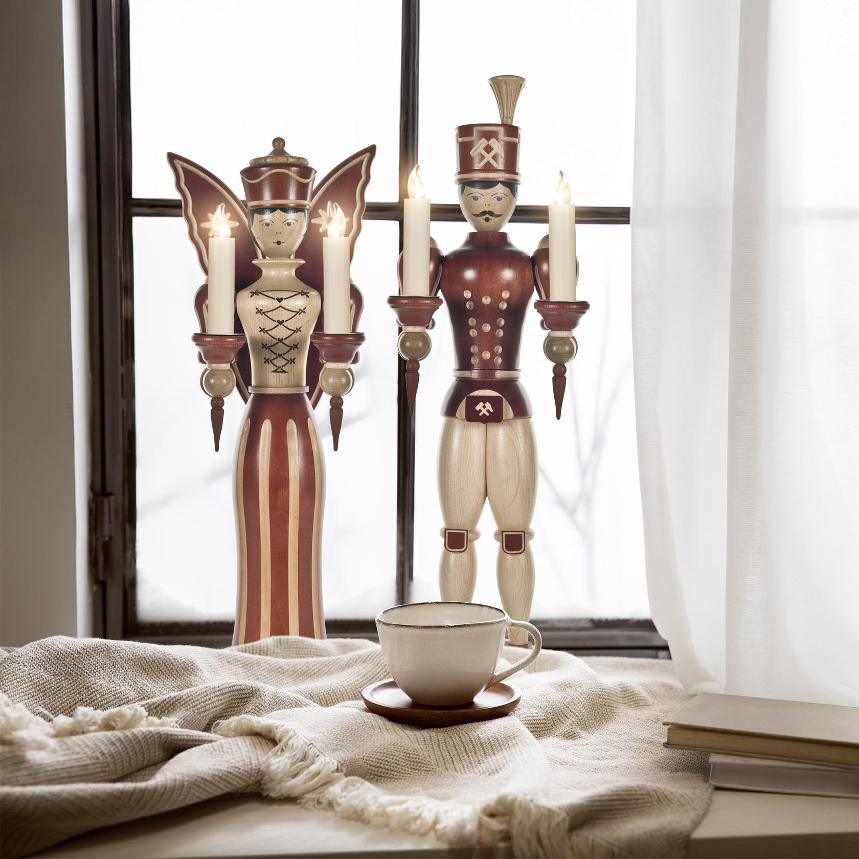 Two wooden figurines, an angel and a miner, holding candles stand by a window. The angel has red and white wings, the miner is in a red cap and uniform.