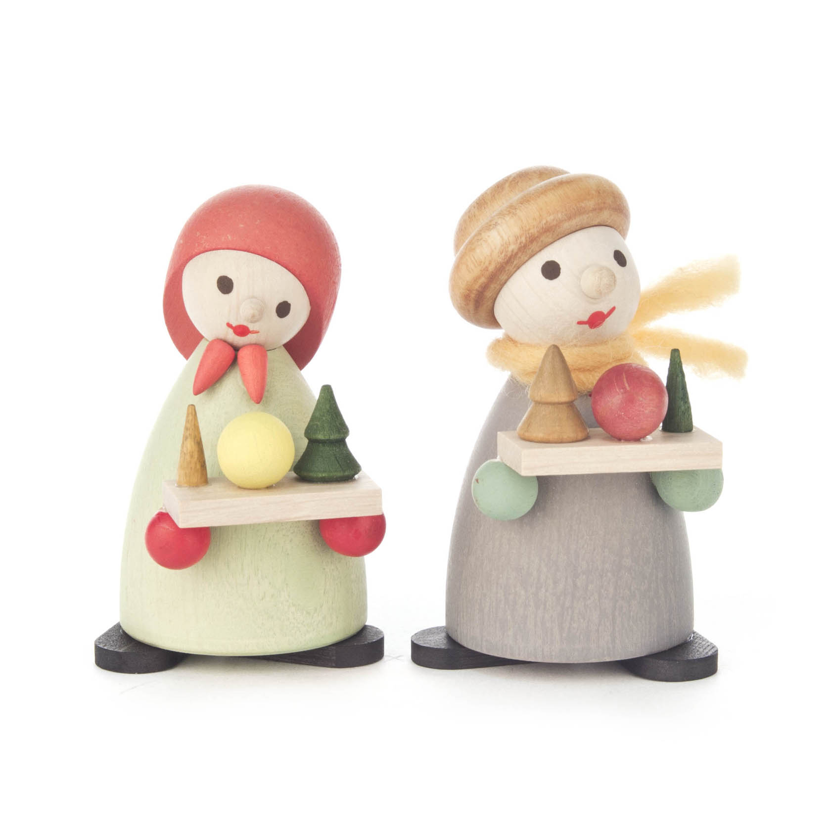 Two modern wooden figurines of children, each holding a wooden tray with colorful miniature trees and ornaments. One wears a red scarf, the other a brown hat.
