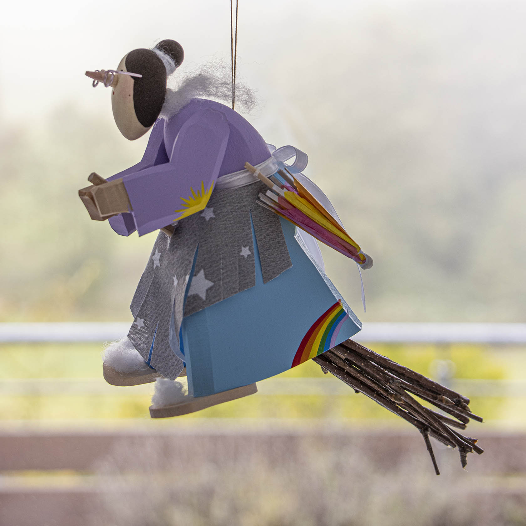 A handmade weather witch figurine with a purple jacket and a star-patterned grey skirt, holding a small umbrella on its back and sitting on a broomstick.